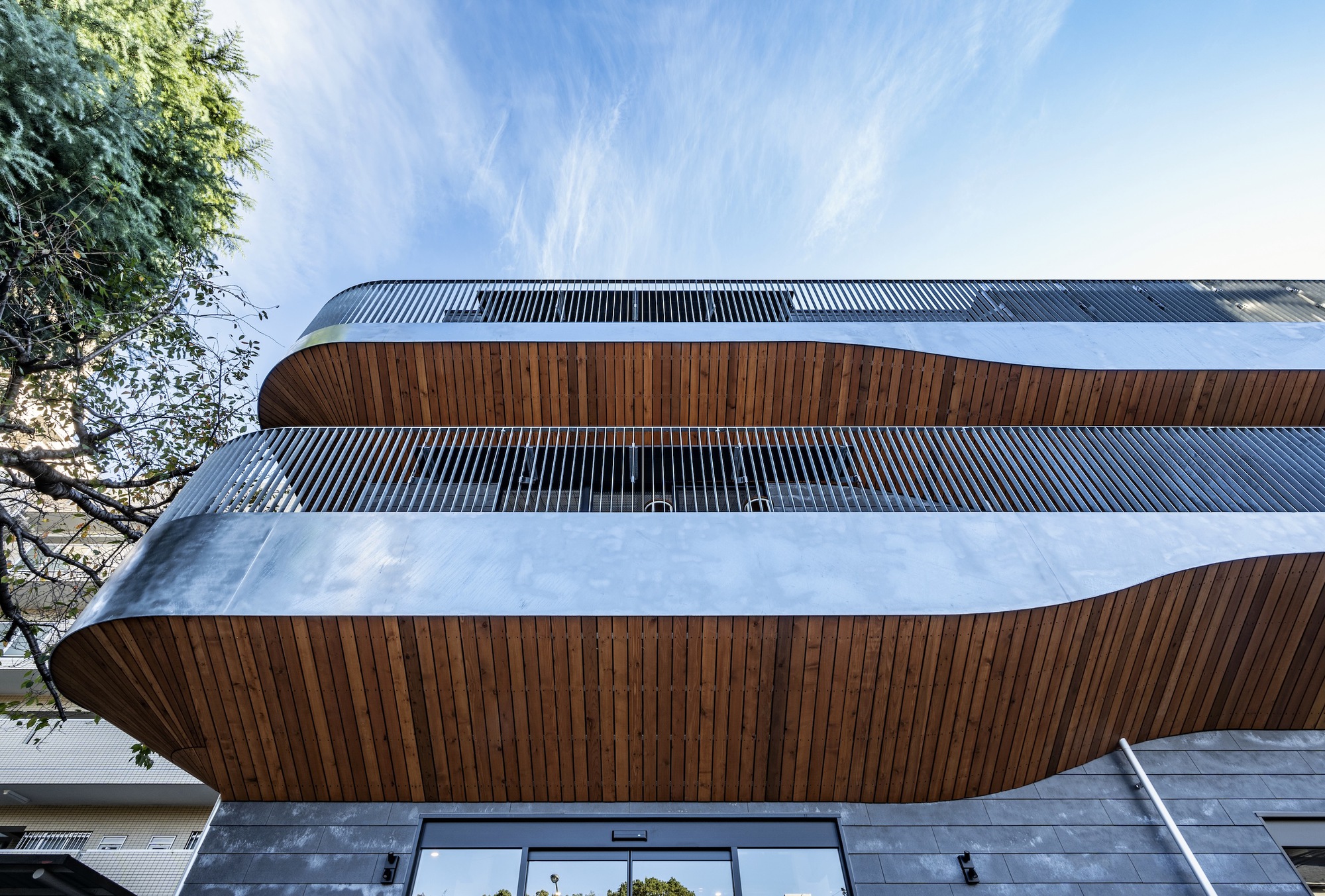 Street view of the curved facade with alternating wood and vertical metal slat bands beneath wispy clouds