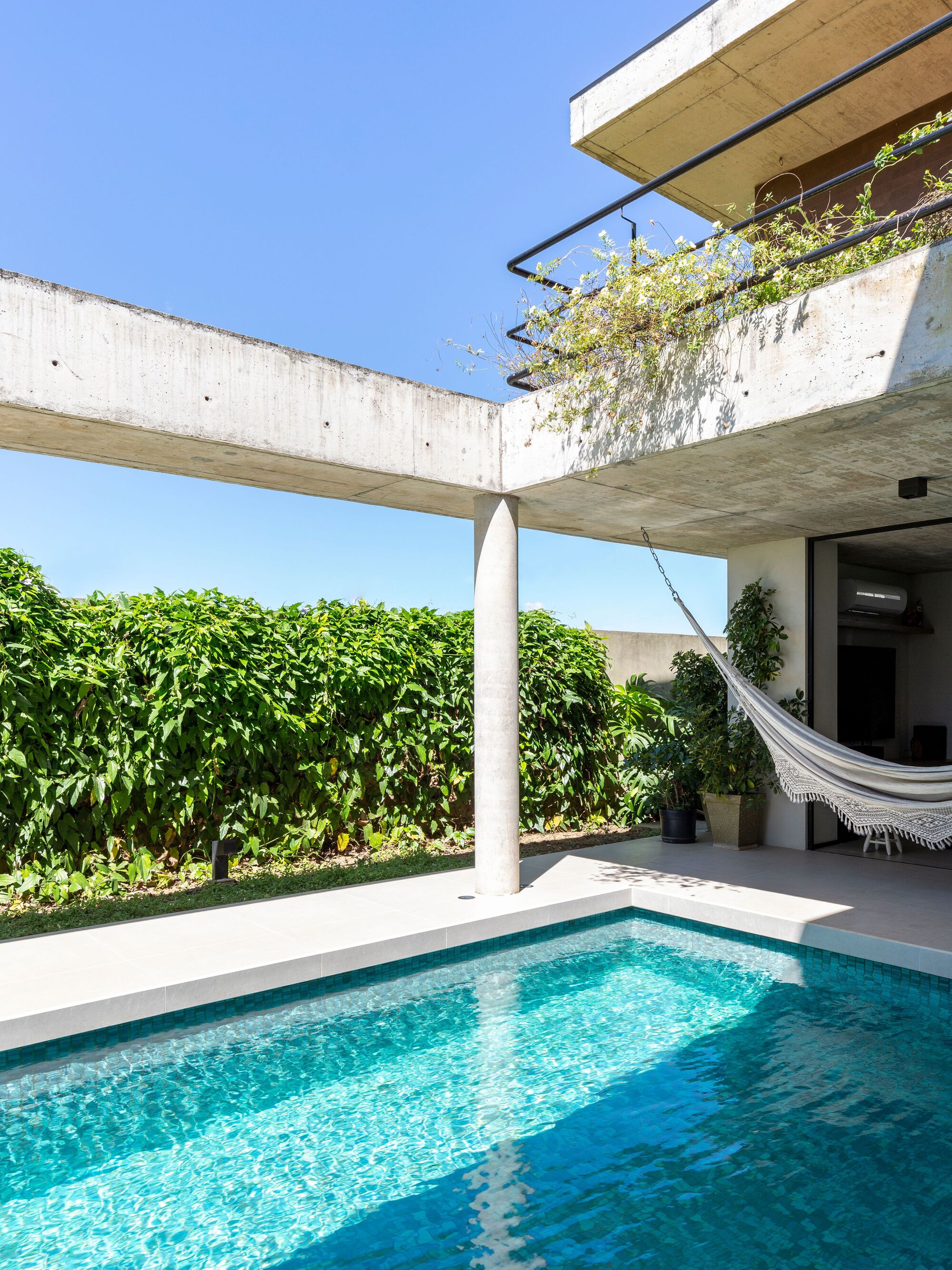 Swimming pool bordered by green hedge wall beneath concrete beam and upper-level planted terrace in daylight