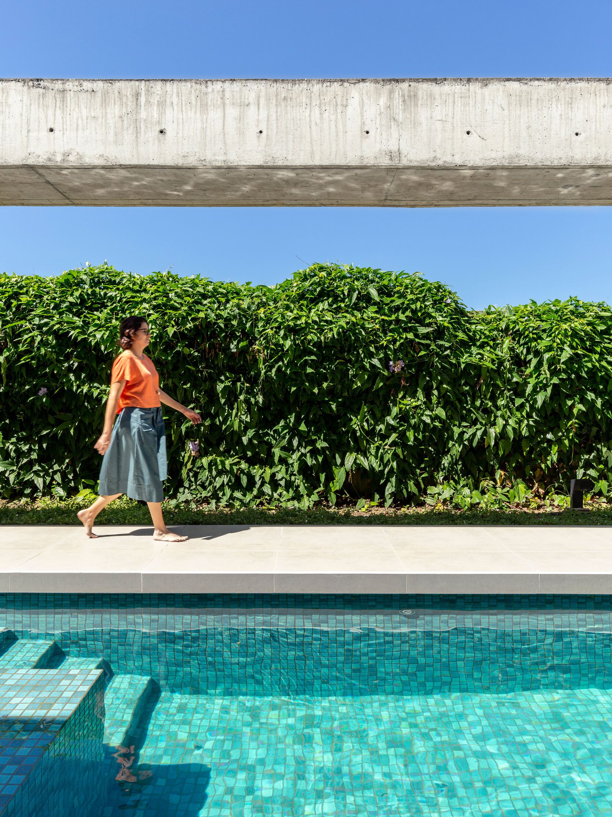 Pool deck with concrete beam overhead, dense hedge boundary, and person in orange shirt walking past