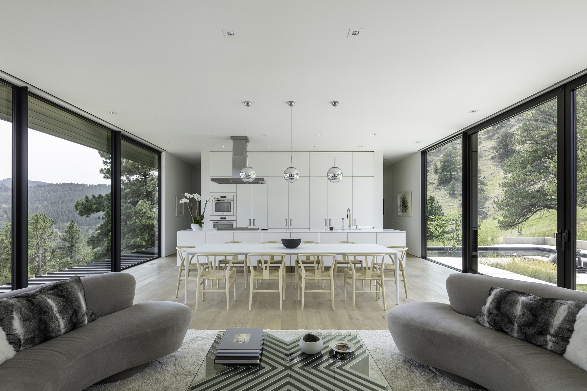 Open-plan kitchen and dining area flanked by floor-to-ceiling glazing with views to forested hills