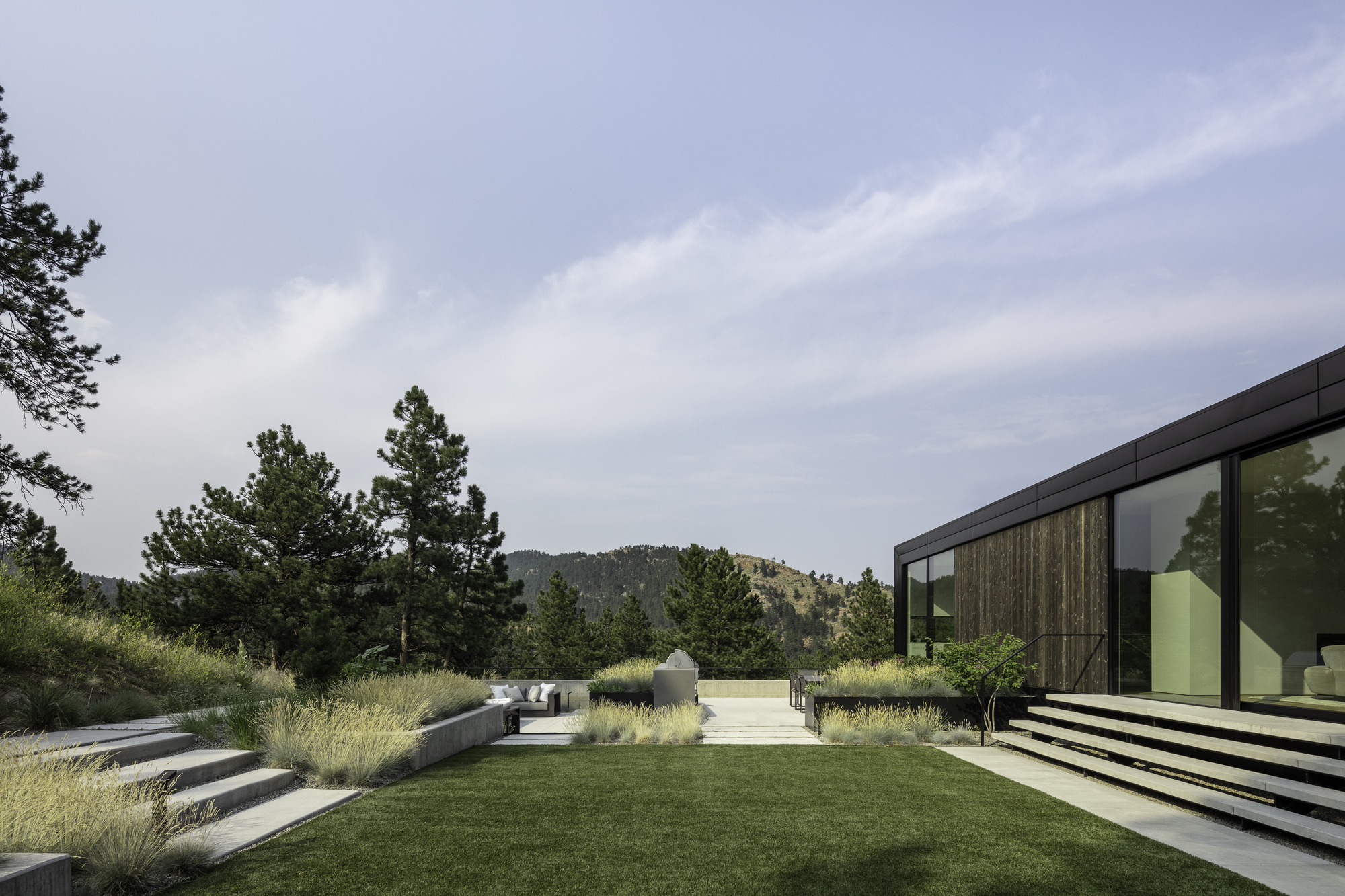 Terraced lawn and gravel pathway extend through native grasses toward forested hills beyond the glass facade