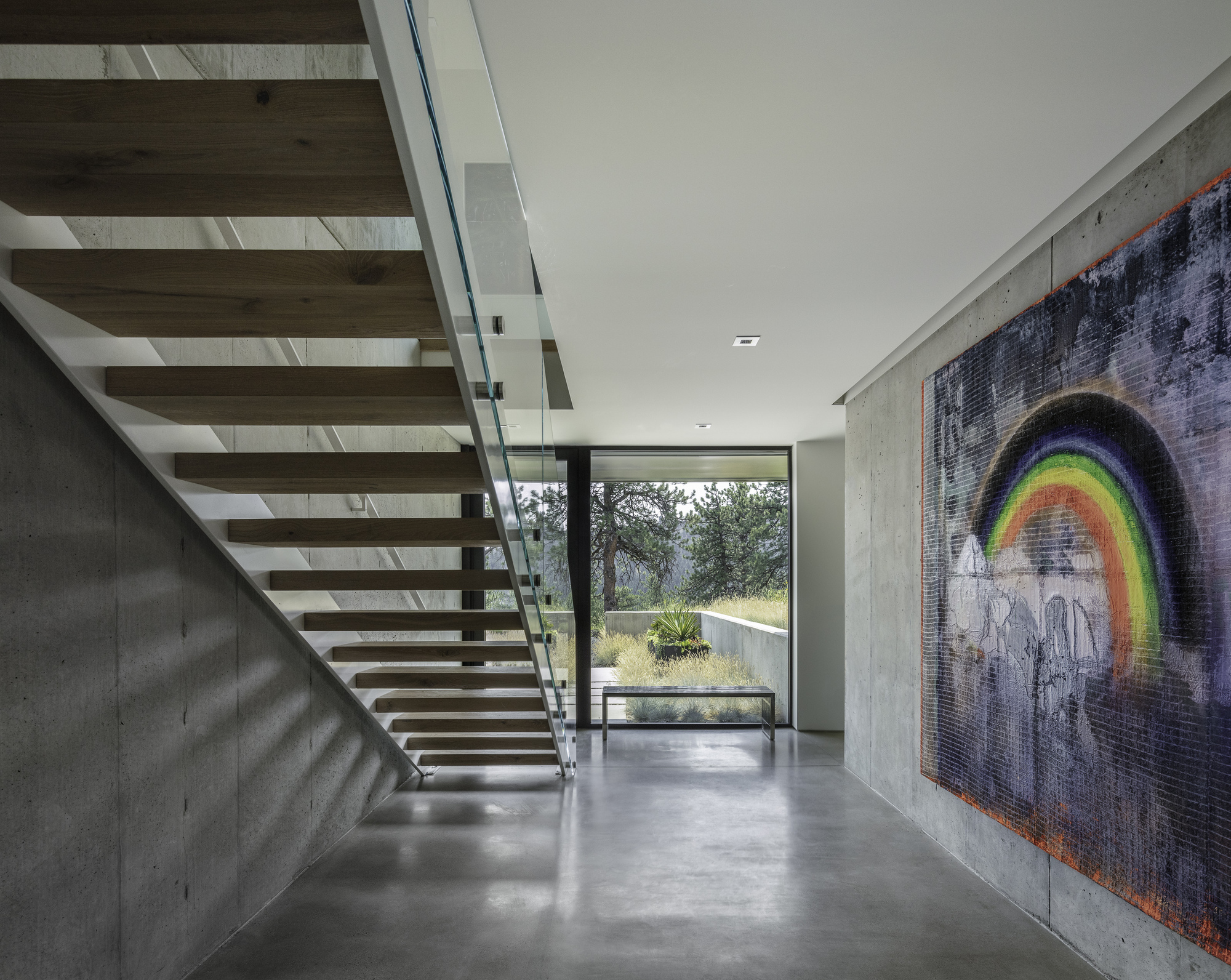 Open tread timber staircase with glass balustrade rises beside a concrete wall displaying a rainbow tapestry