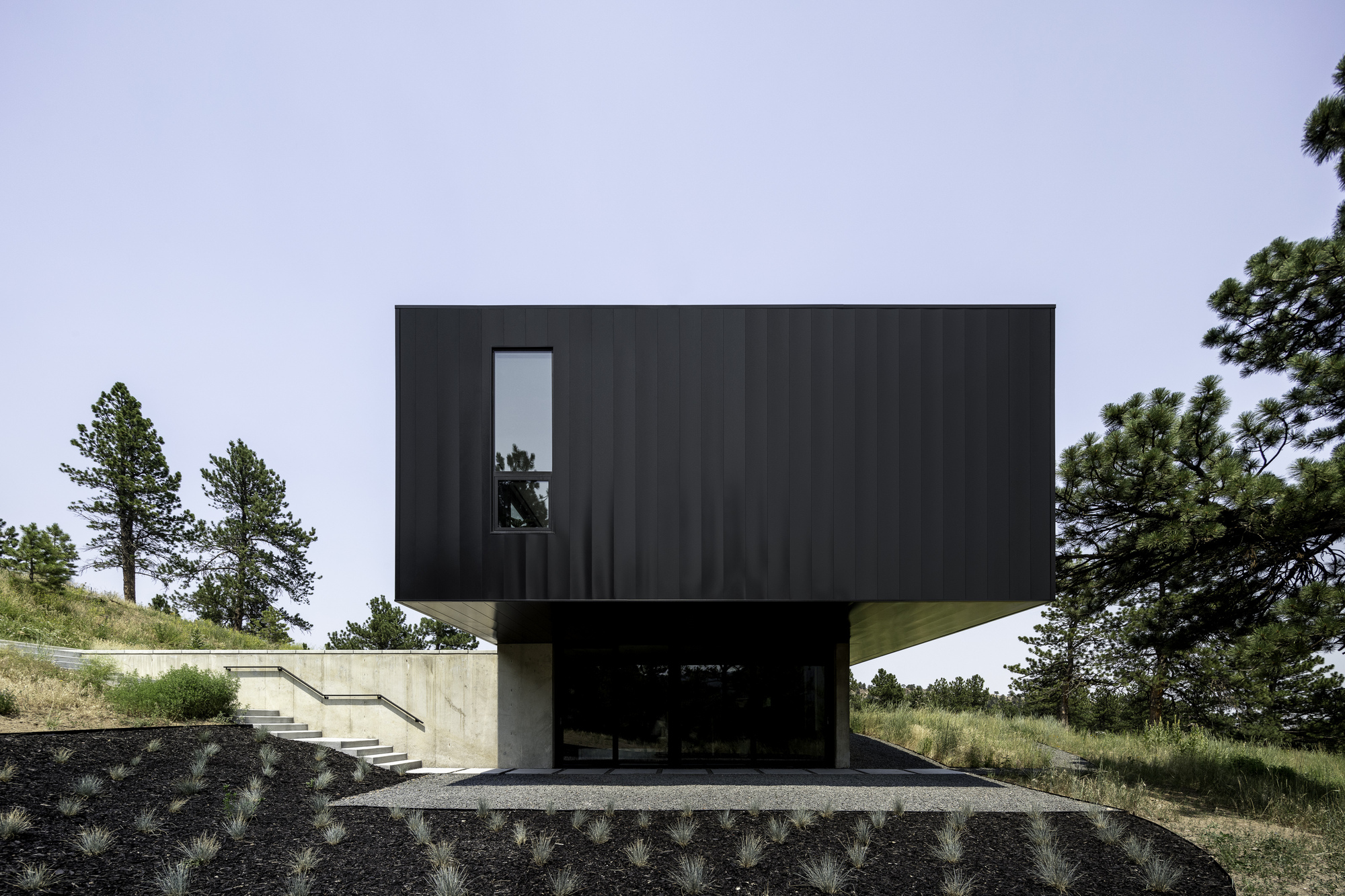 Dark metal clad volume perches above a concrete plinth with open carport cut into the hillside