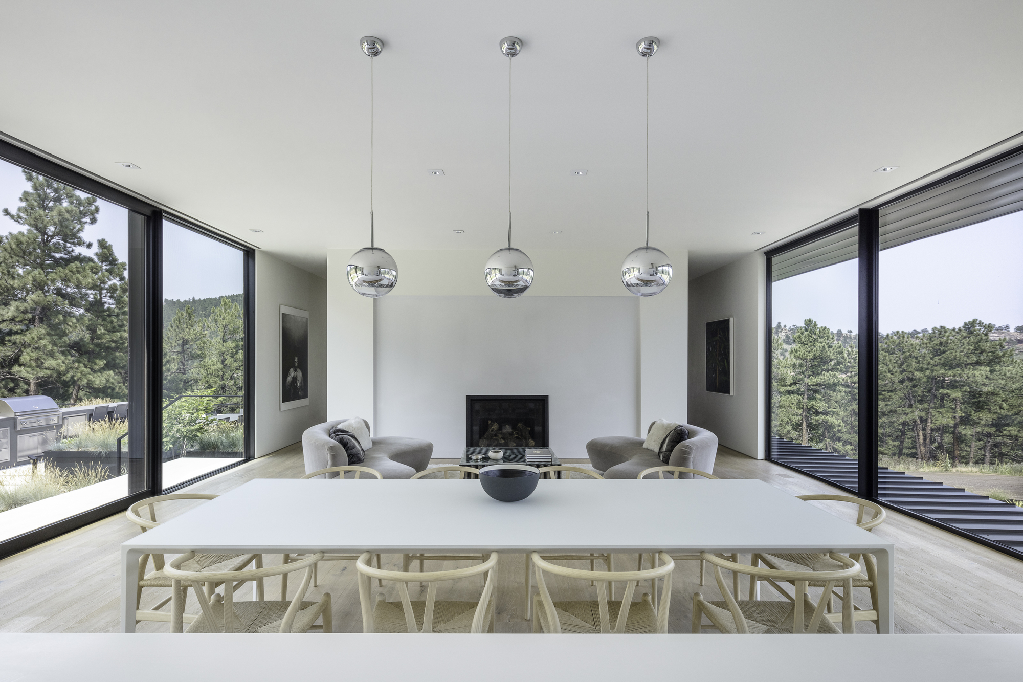 White dining table with wishbone chairs beneath three globe pendant lights and symmetrical corner windows