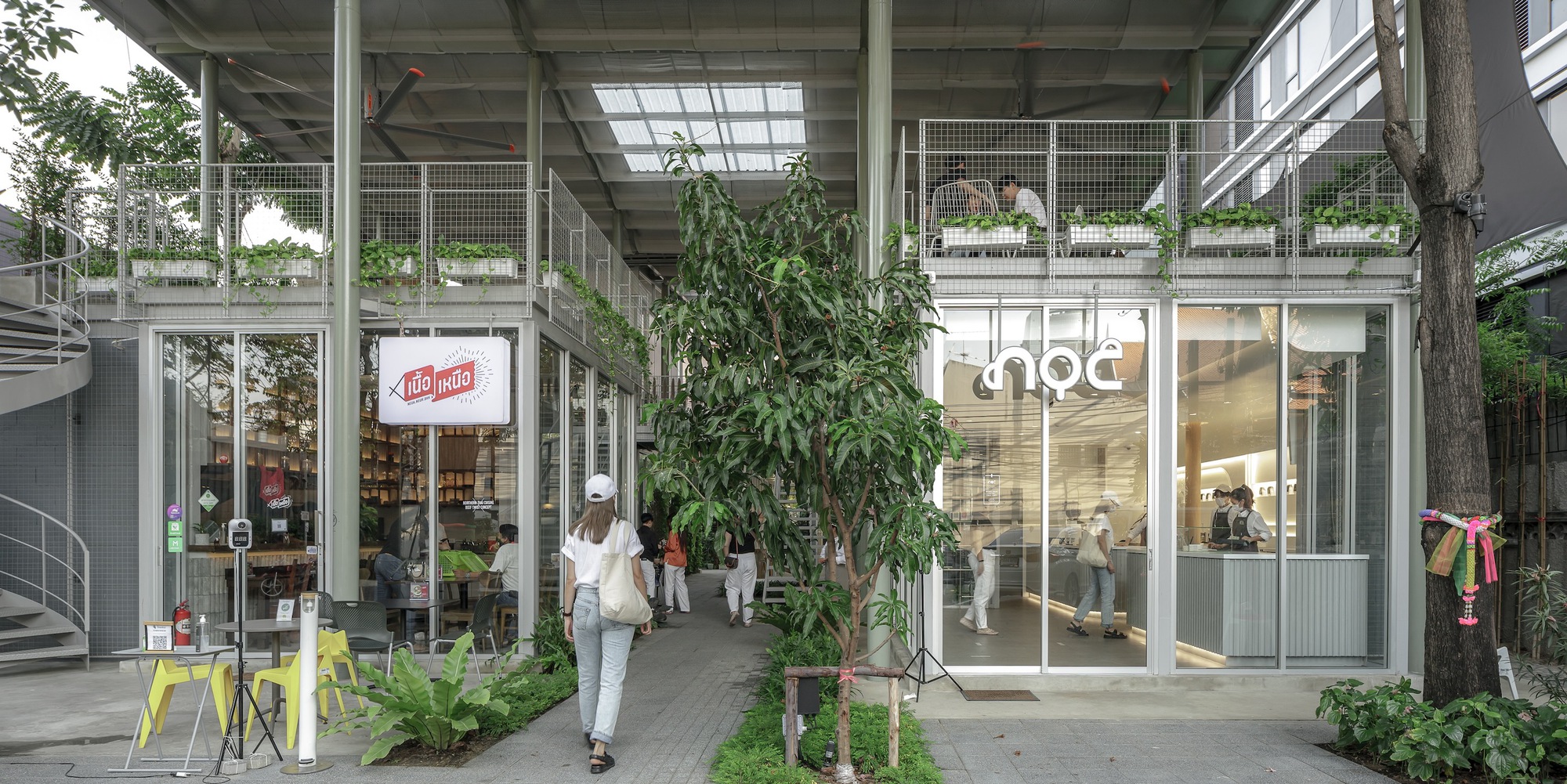 Ground-level passage with glass storefronts and mesh railings as a visitor walks through planted greenery