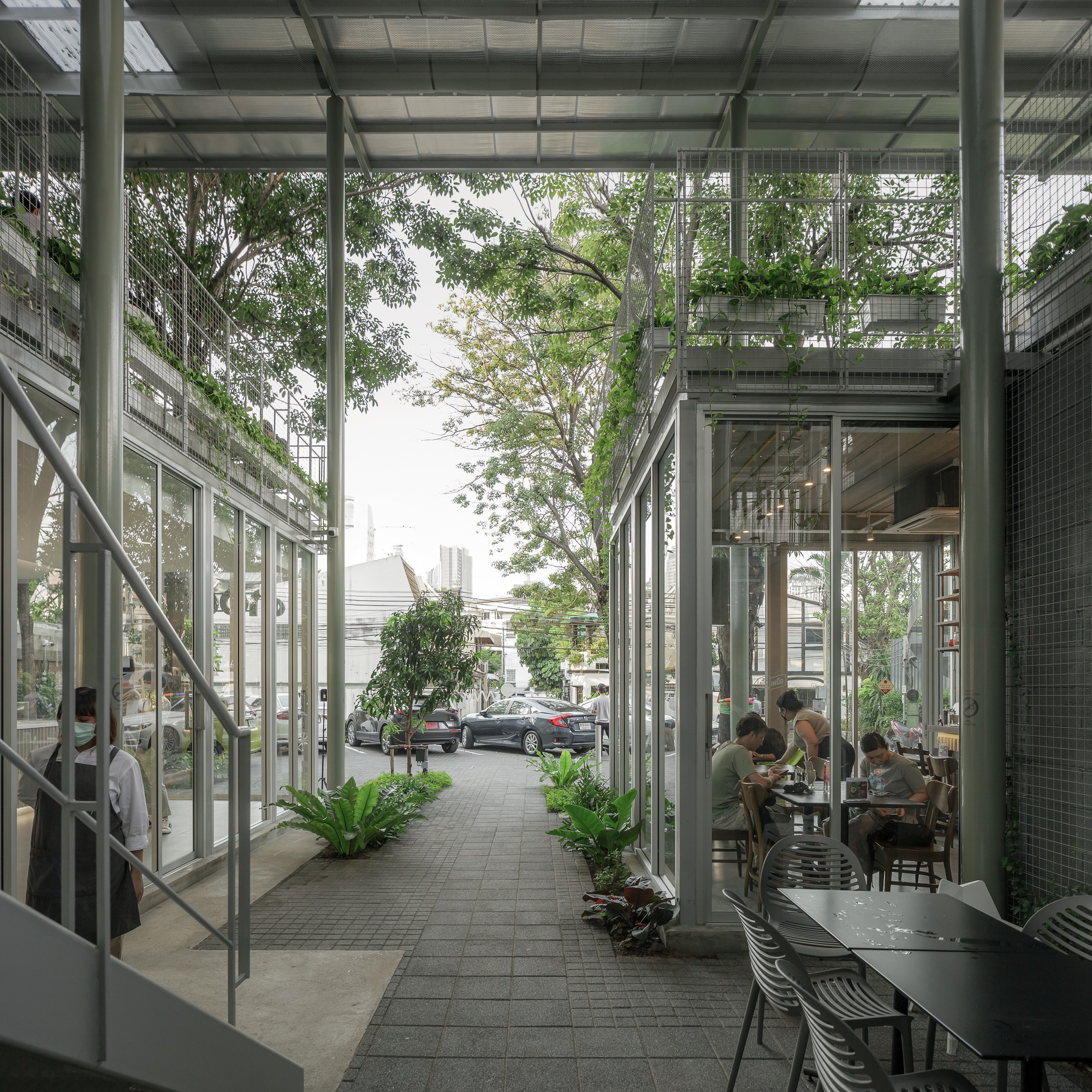 Pedestrian lane framed by steel columns and glazing with planters and diners visible through the cafe windows