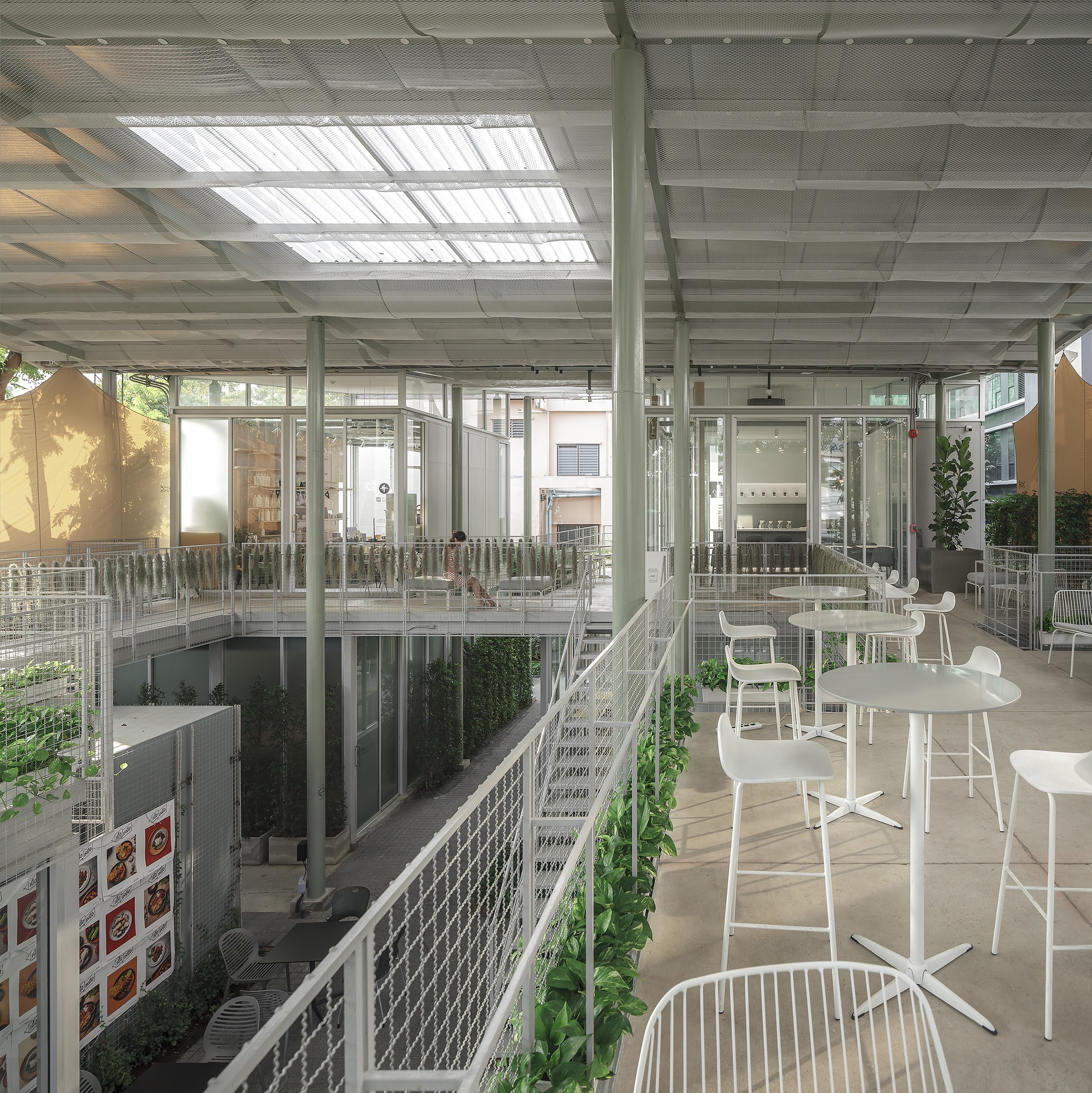Interior terrace with white furniture and mesh railings overlooking planted voids under the translucent ceiling grid