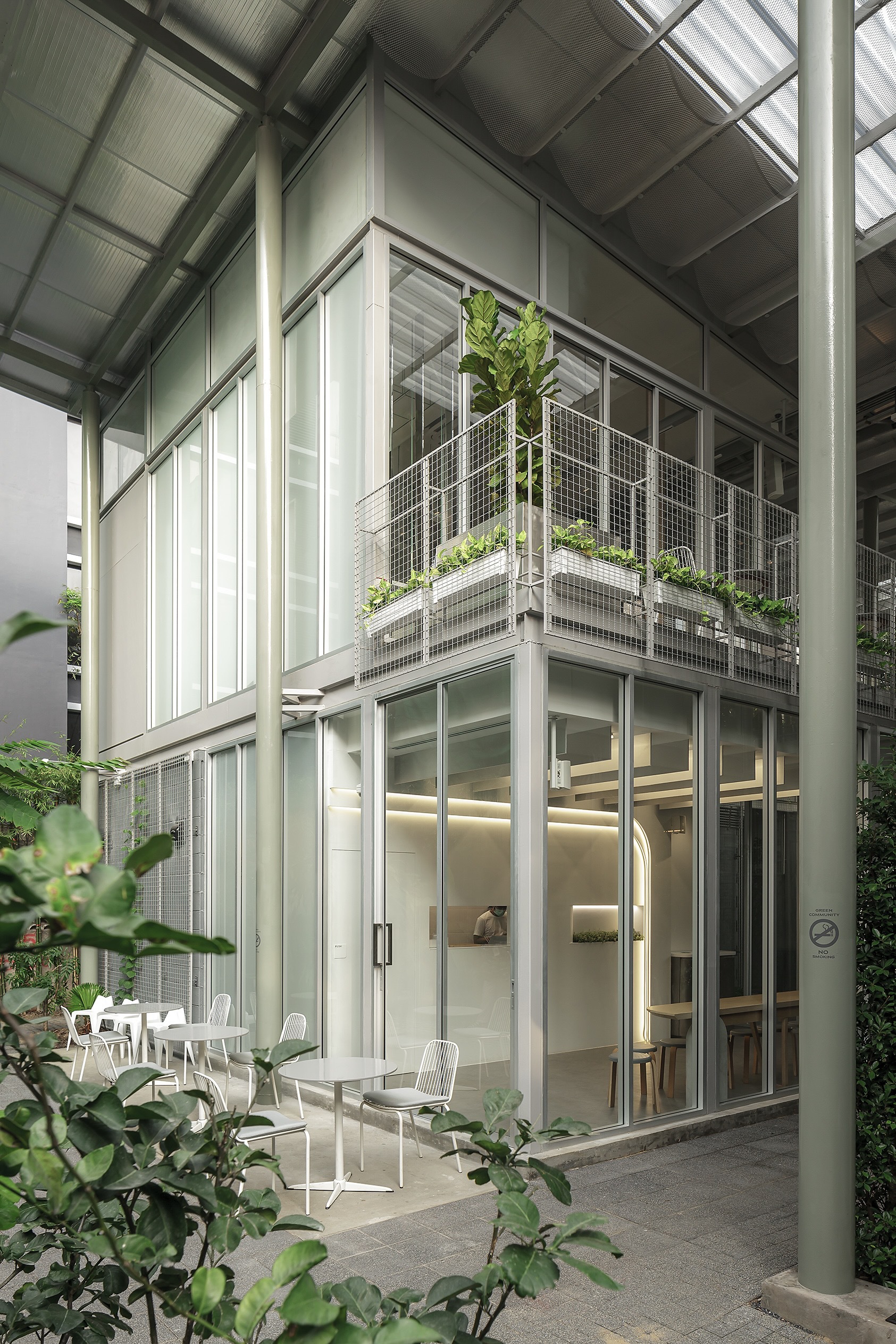 Two-storey glazed facade with planted wire mesh balcony and white metal chairs on the ground level