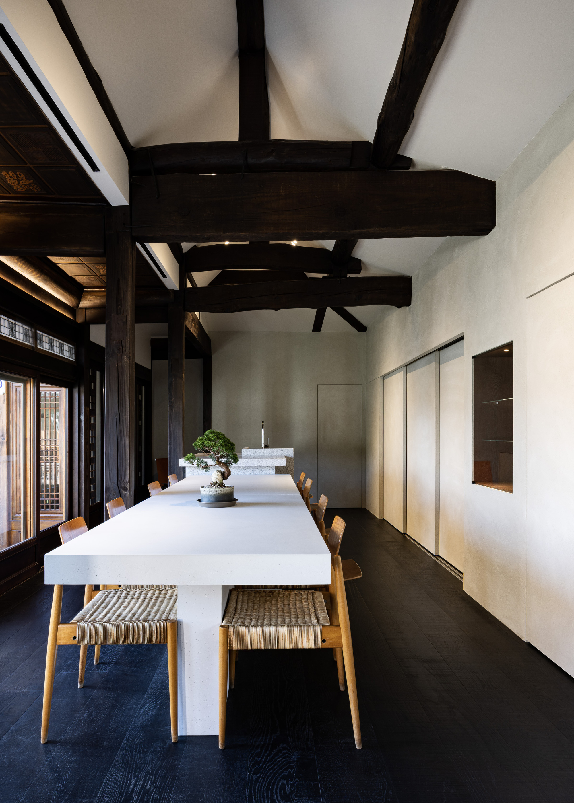 Dining room with exposed dark timber beams above a white table and wooden chairs on dark flooring