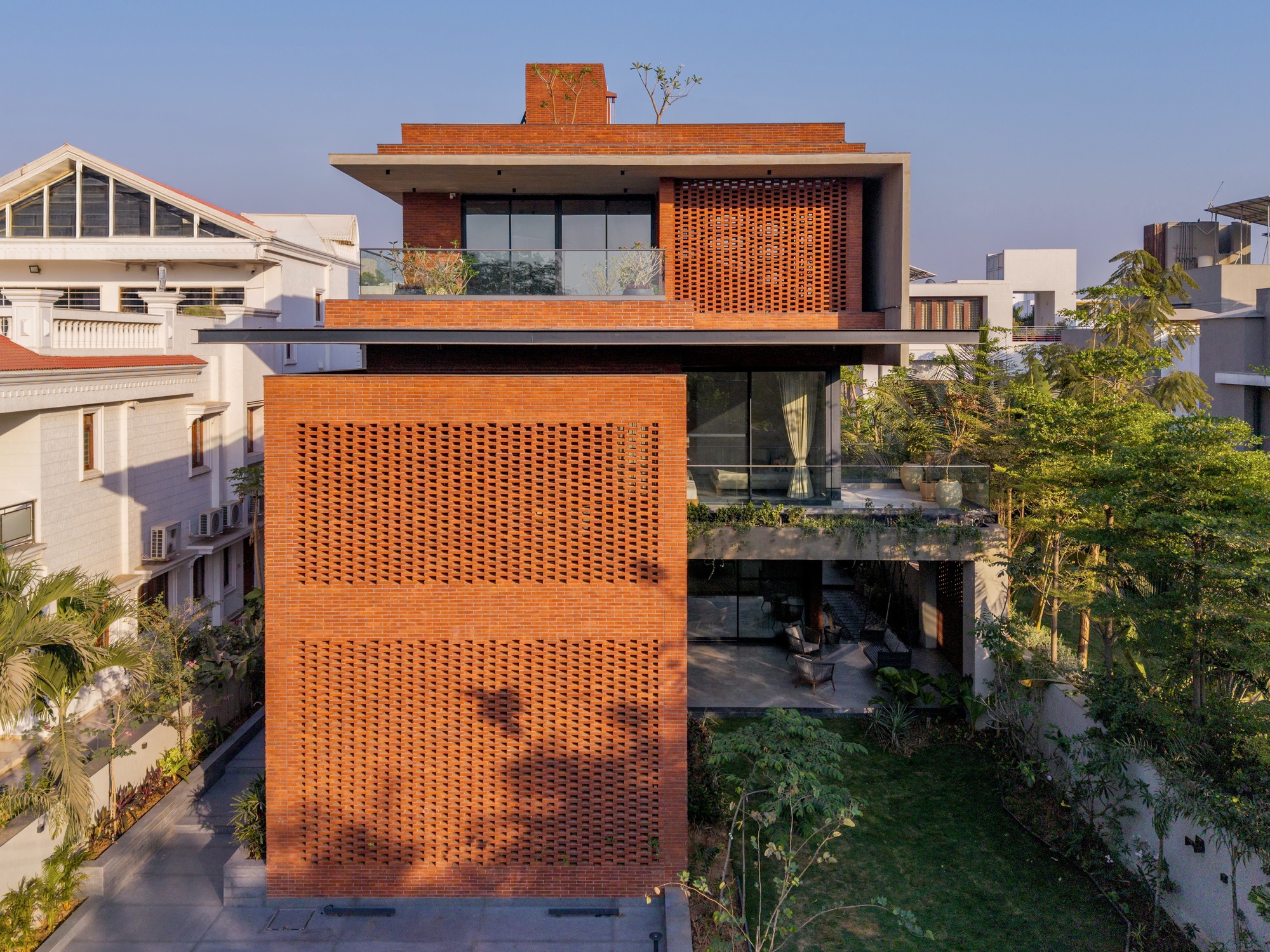 Three-story brick facade with perforated screen walls and rooftop terrace amid neighboring residential structures
