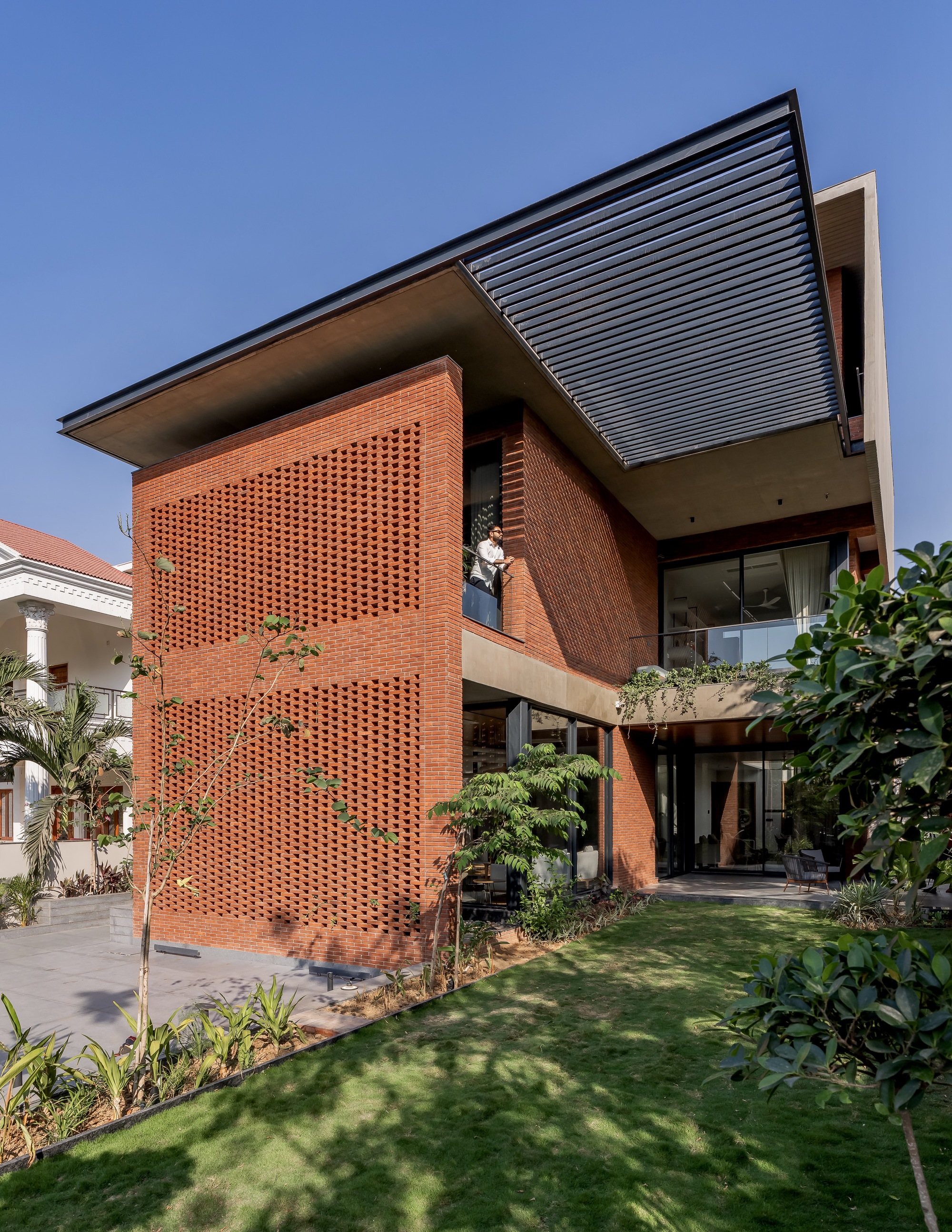 Garden view of perforated red brick facade and cantilevered metal roof under clear blue sky