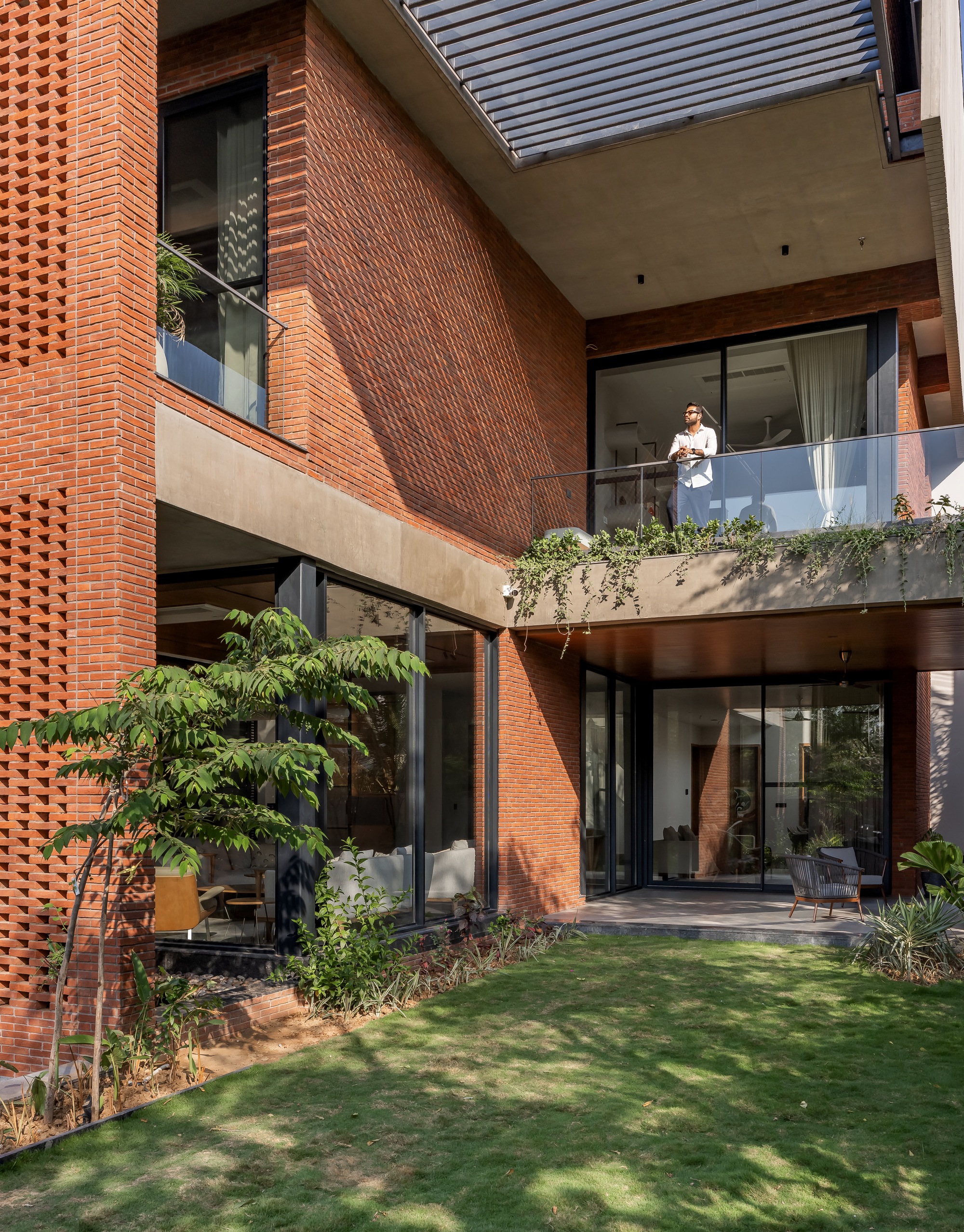 Perforated brick facade with planted balcony where a resident stands overlooking the garden below