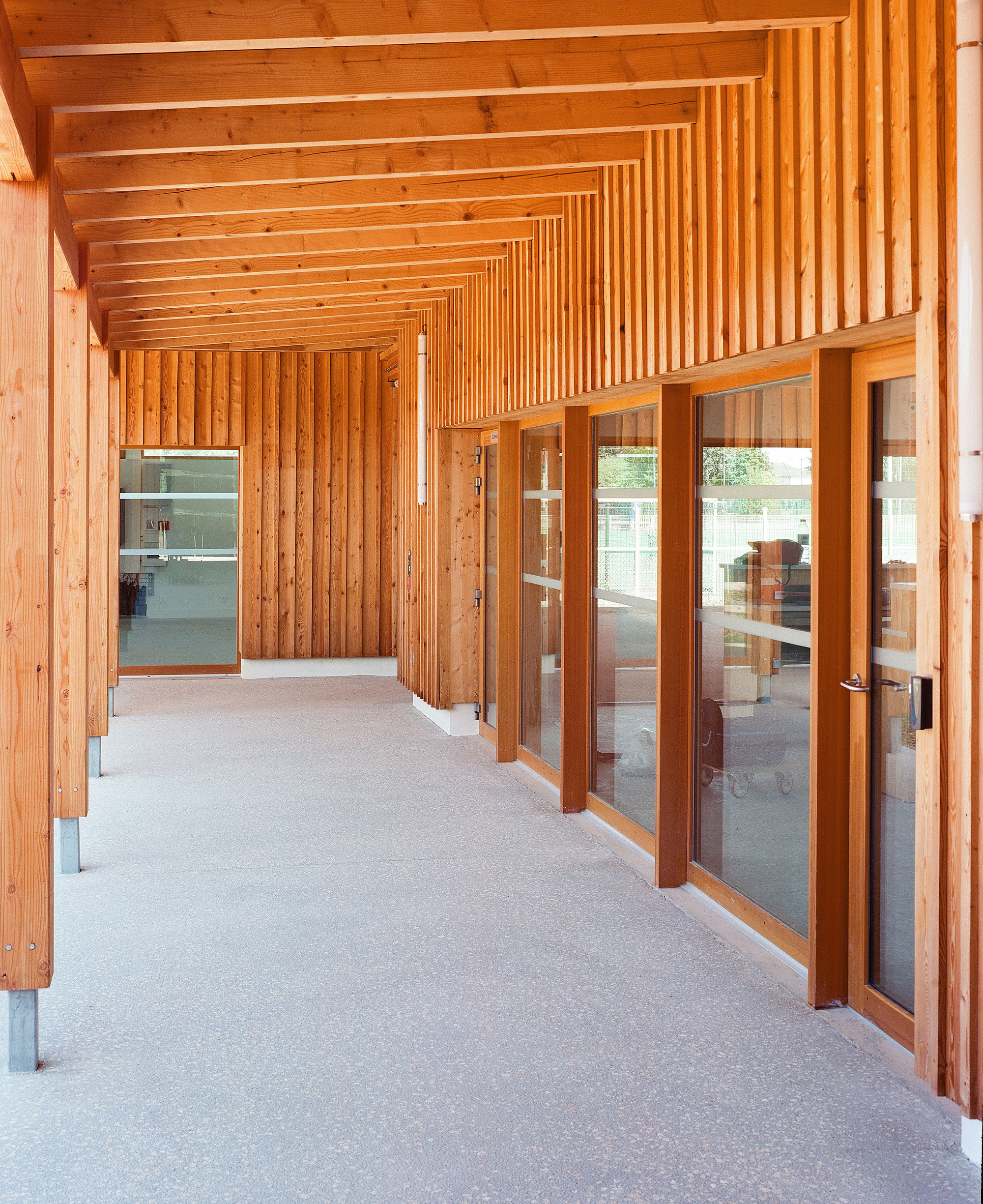 Timber-clad corridor with glazed doors, vertical slat screen, and exposed beam ceiling in daylight