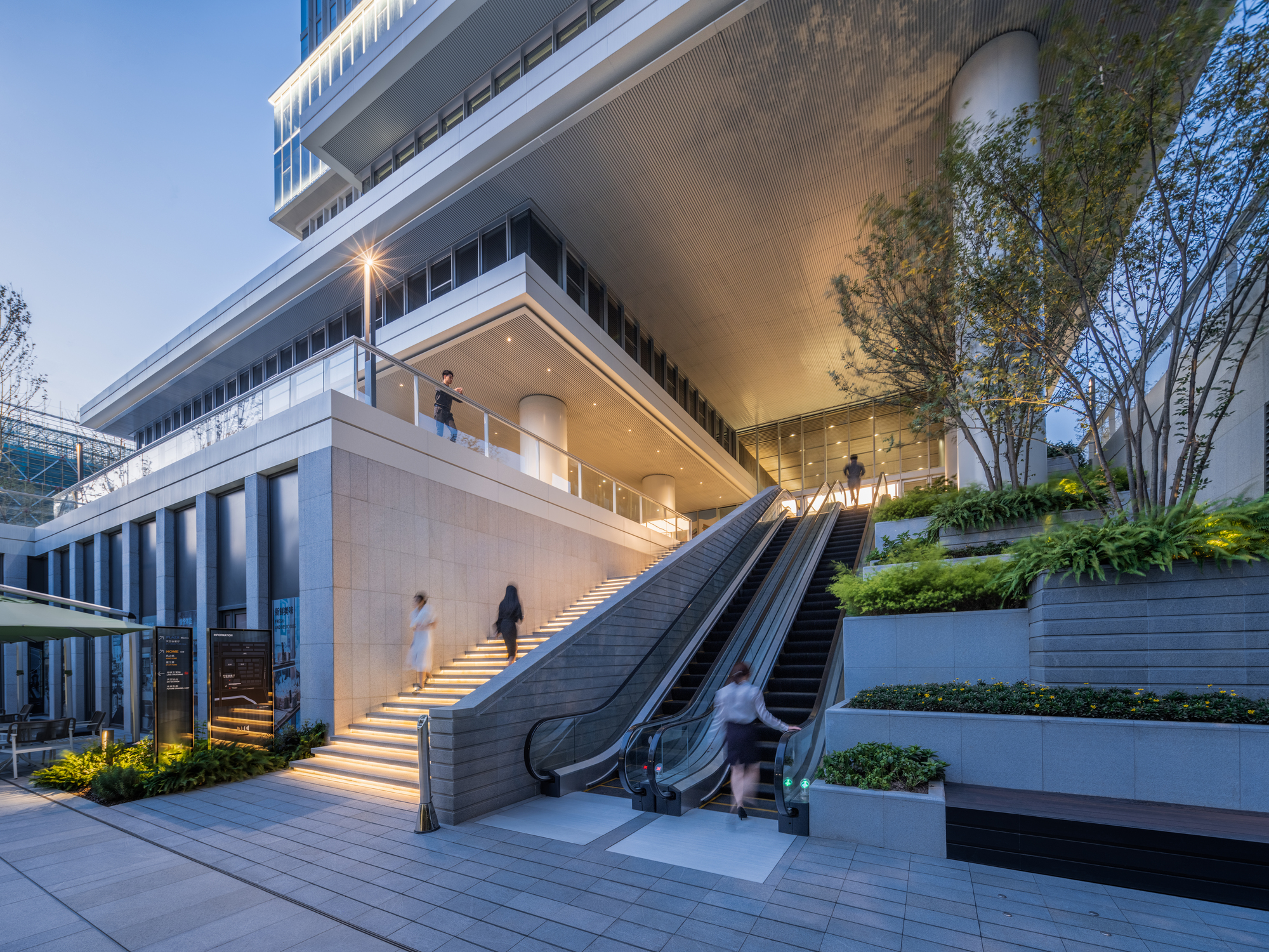 Outdoor staircase and escalator flanked by planted terraces with people ascending at twilight