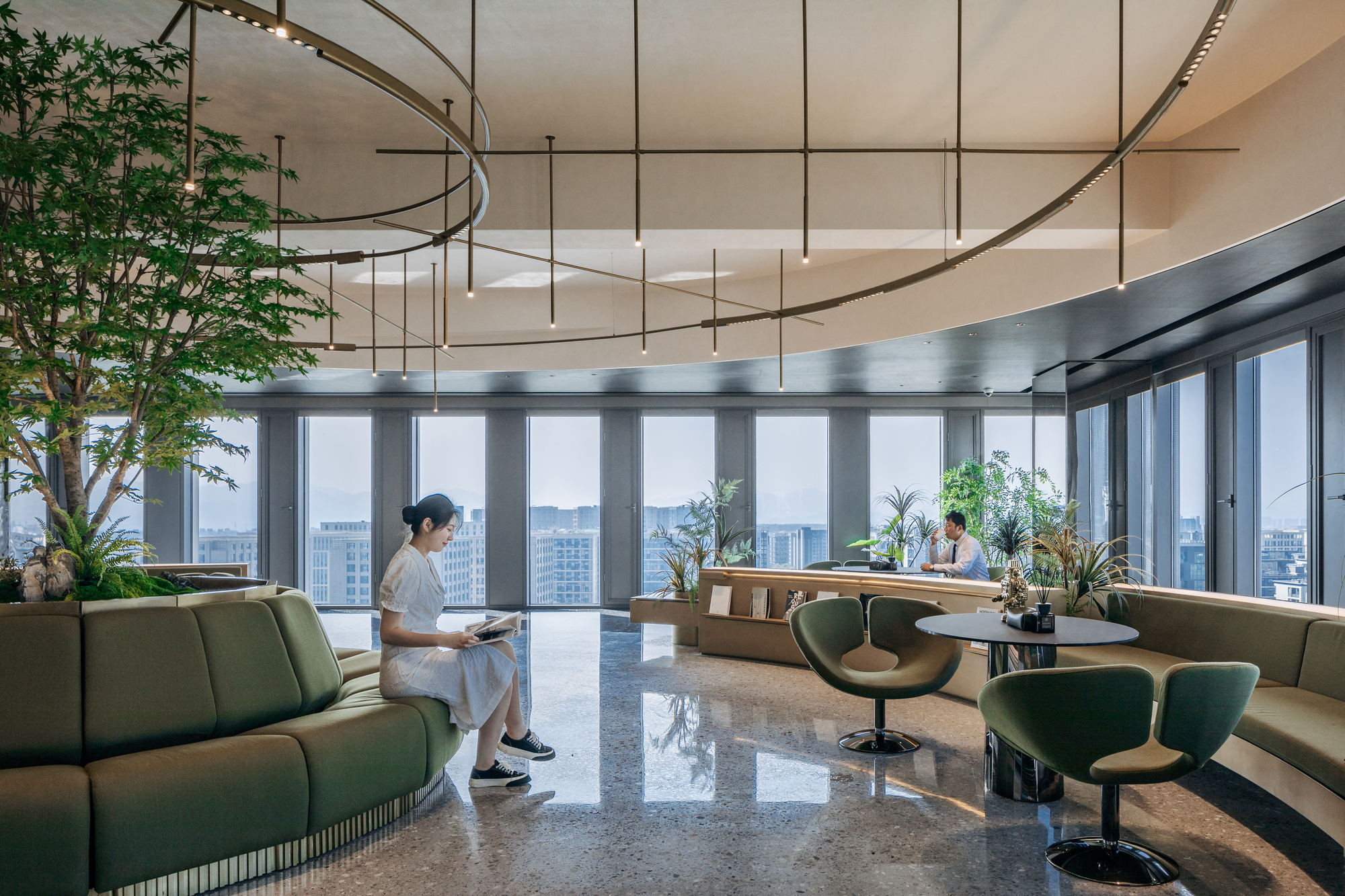 Interior lounge with green seating, terrazzo flooring, suspended circular light fixtures and a person reading