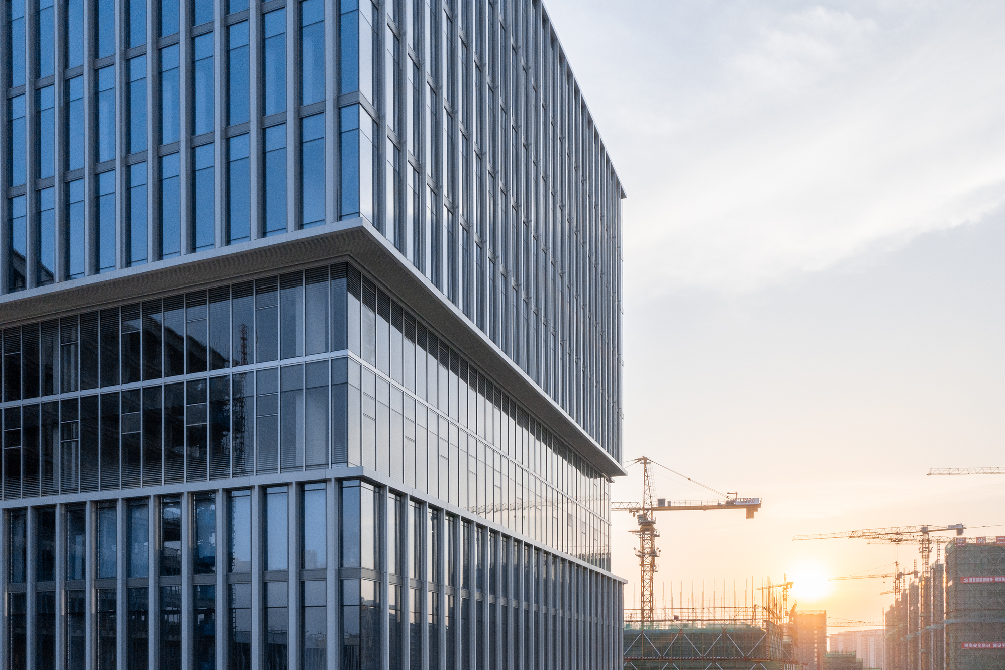 Corner facade with vertical metal fins and recessed glass volumes at sunset with cranes visible
