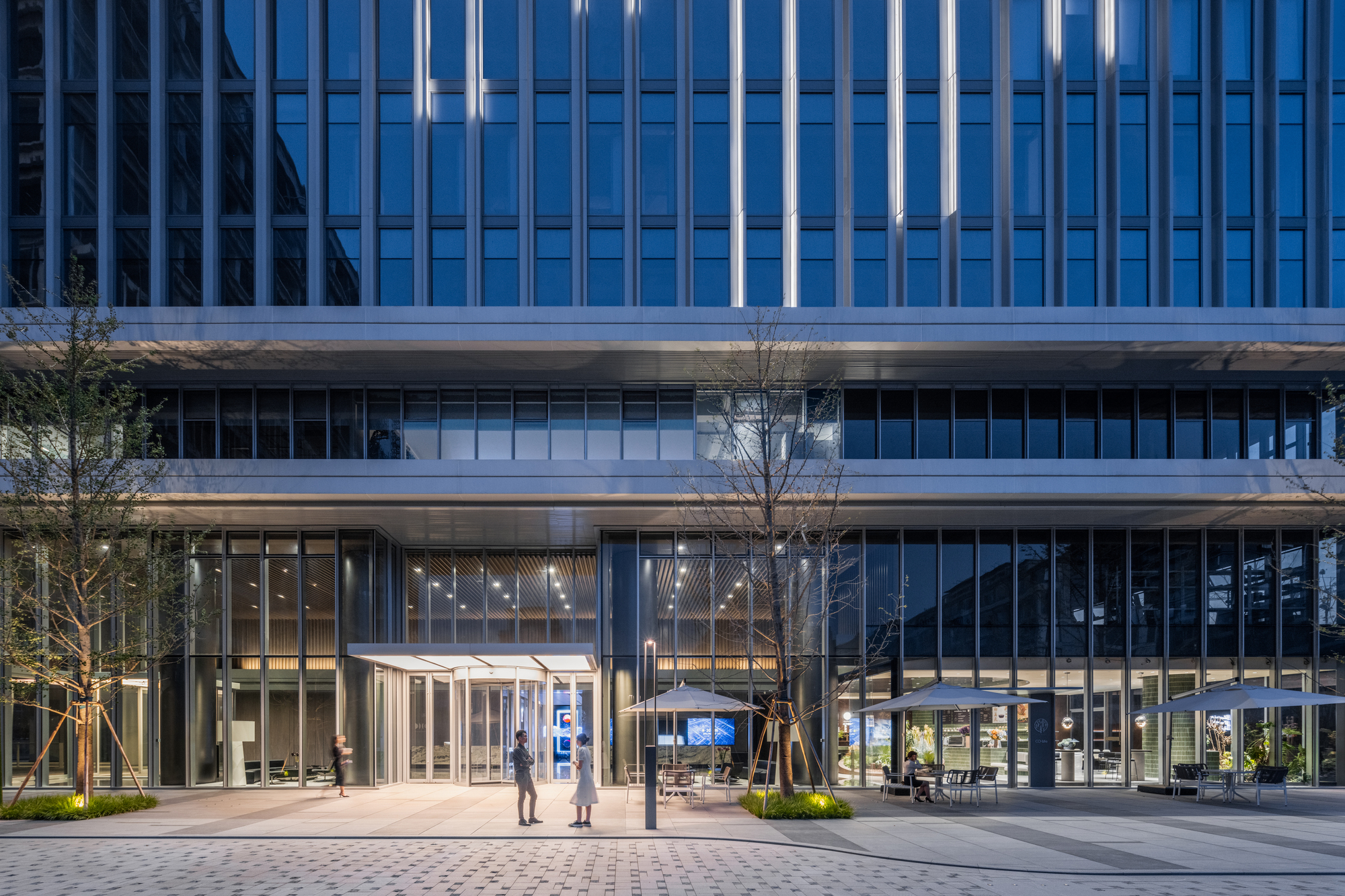 Ground level plaza with glass entrance pavilion, paired figures walking, and young trees at dusk