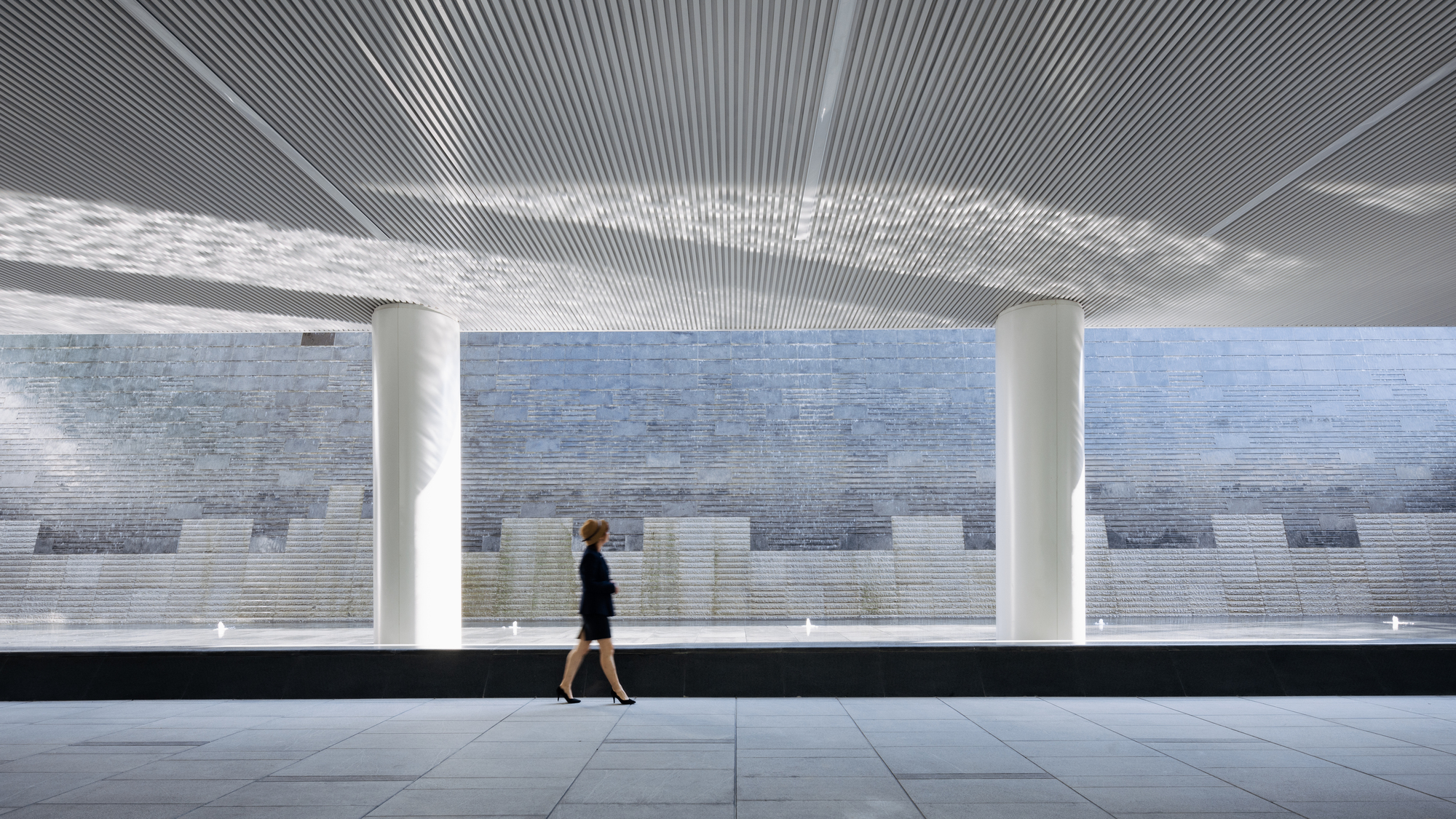 Covered walkway with ribbed ceiling and white columns as a person passes the pixelated tile wall