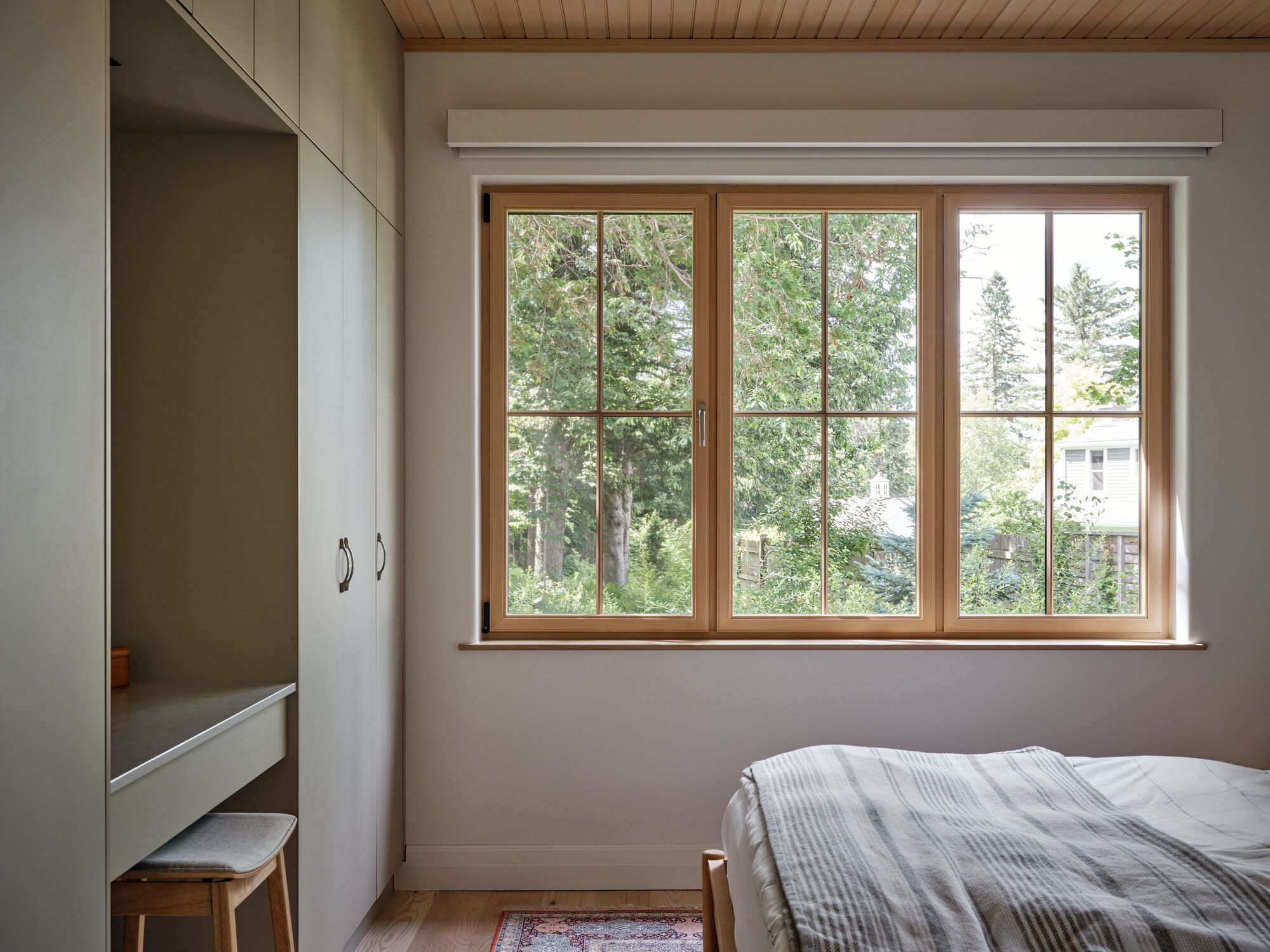 Bedroom with timber-framed casement windows overlooking summer foliage and neighboring houses