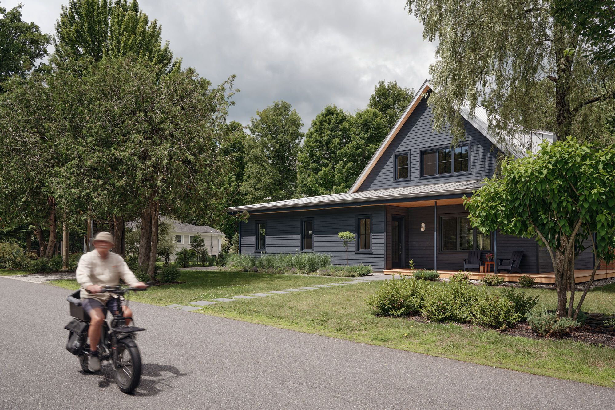 Street view of dark timber-clad gabled house with cyclist passing under cloudy sky