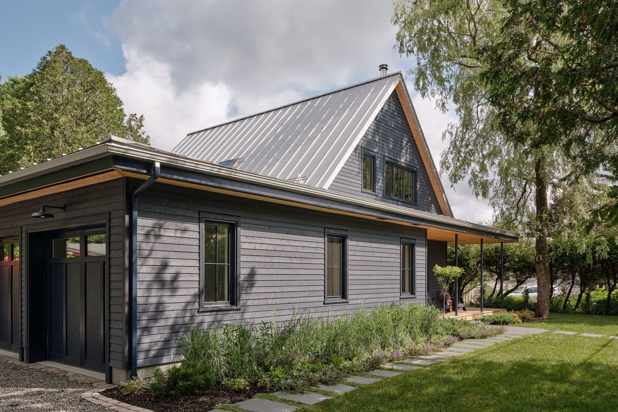 Exterior showing metal standing-seam roof and dark horizontal siding beside planted beds and stone path