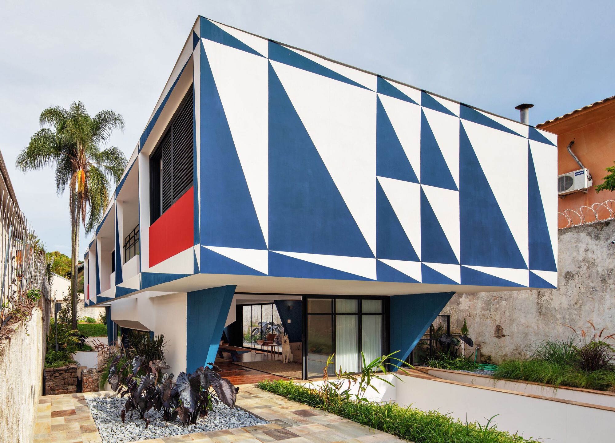 Cantilevered upper volume with blue and white triangular painted facade above courtyard with palm trees