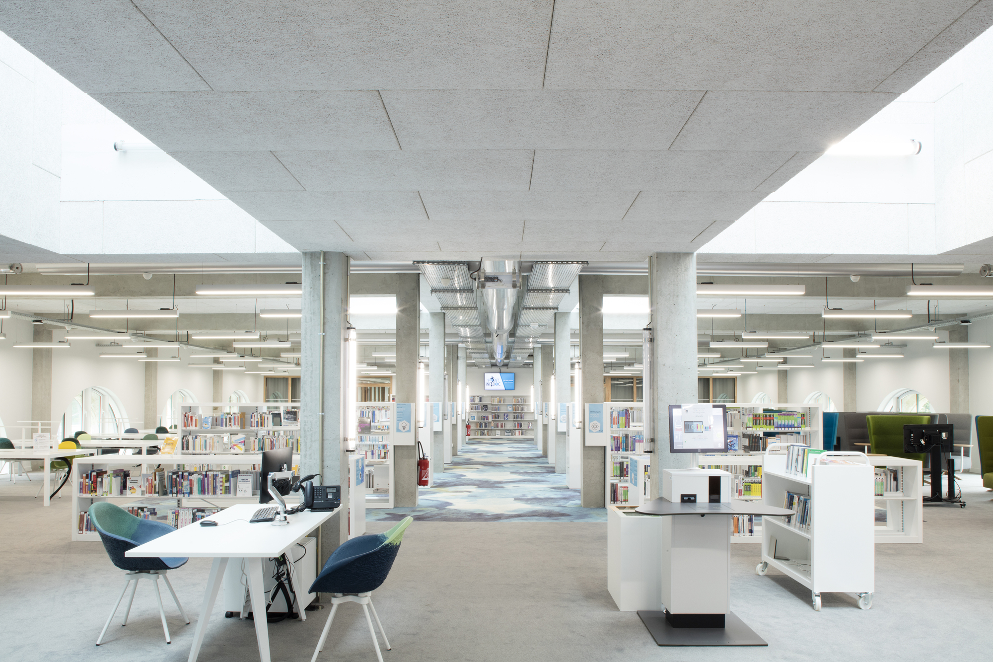 Library interior with concrete columns, exposed ductwork and white bookshelves under a coffered concrete ceiling