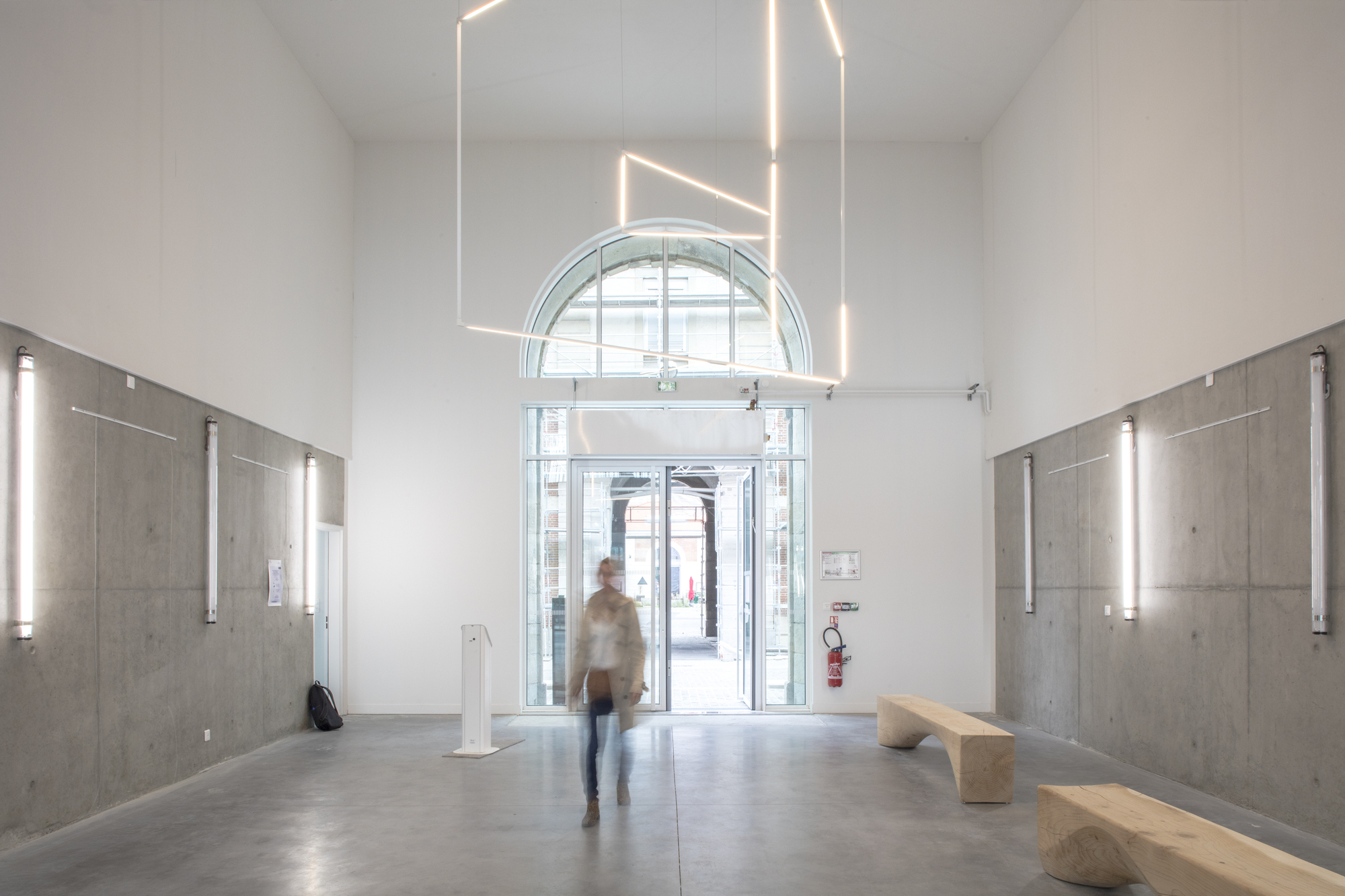 Entry hall with concrete wall panels, linear pendant lighting and a figure walking toward the arched doorway