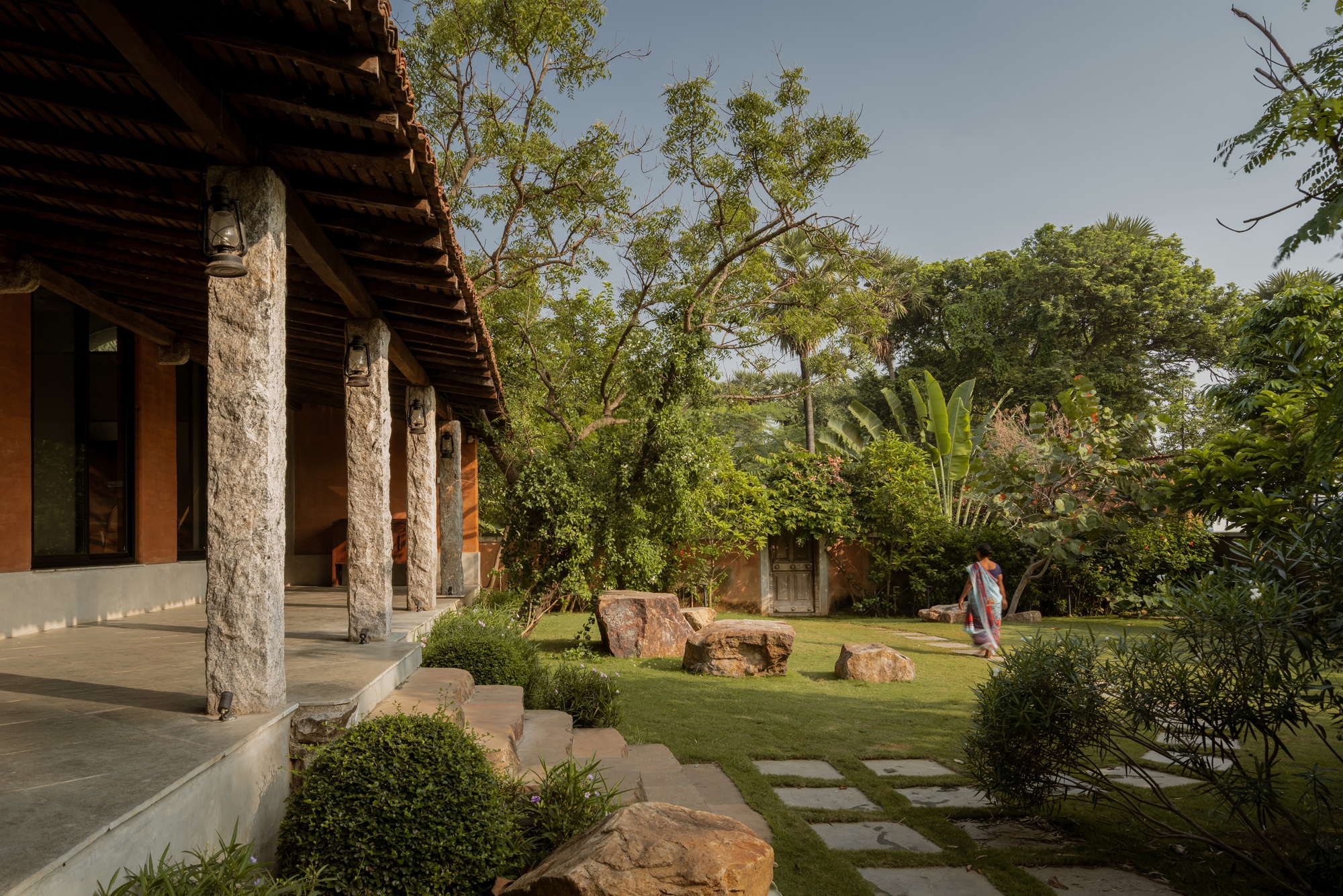 Covered veranda with stone columns and timber roof overlooking a garden with stepping stones and a visitor walking