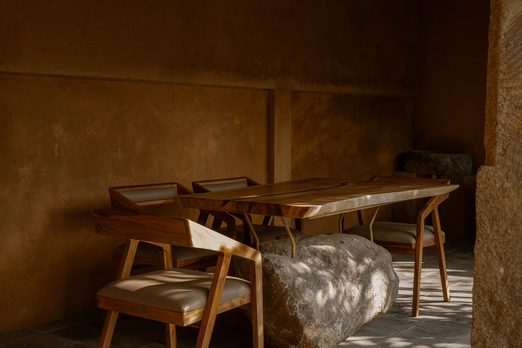 Timber dining table with boulder base and chairs in warm sunlight against textured earthen walls