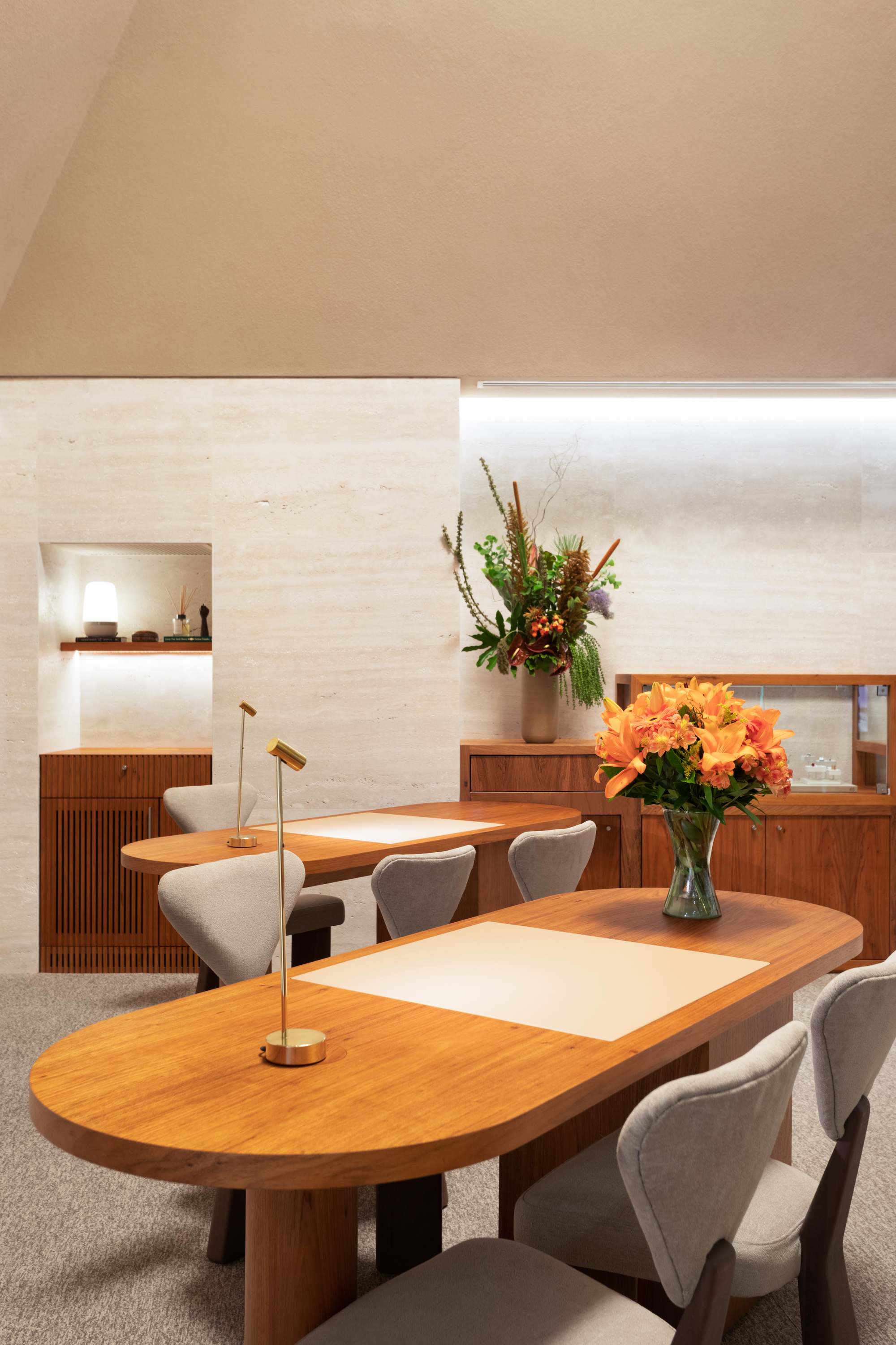 Timber tables with fresh floral arrangements beneath a linear skylight and travertine wall niche