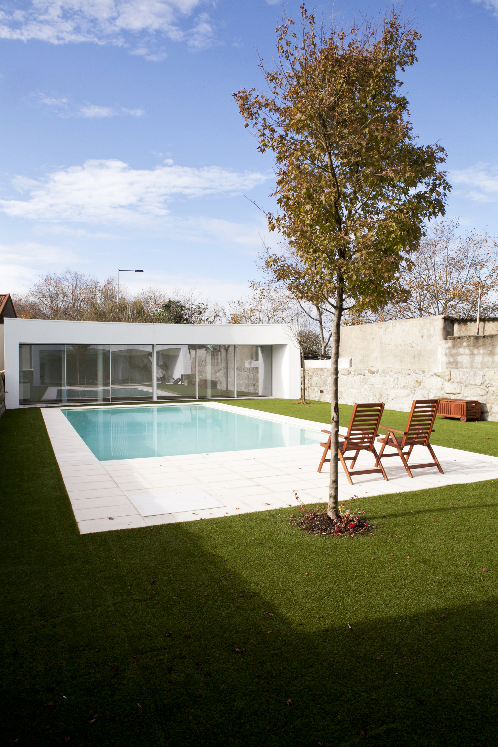 Rear lawn with planted tree, poolside deck and low white pavilion under autumn sky