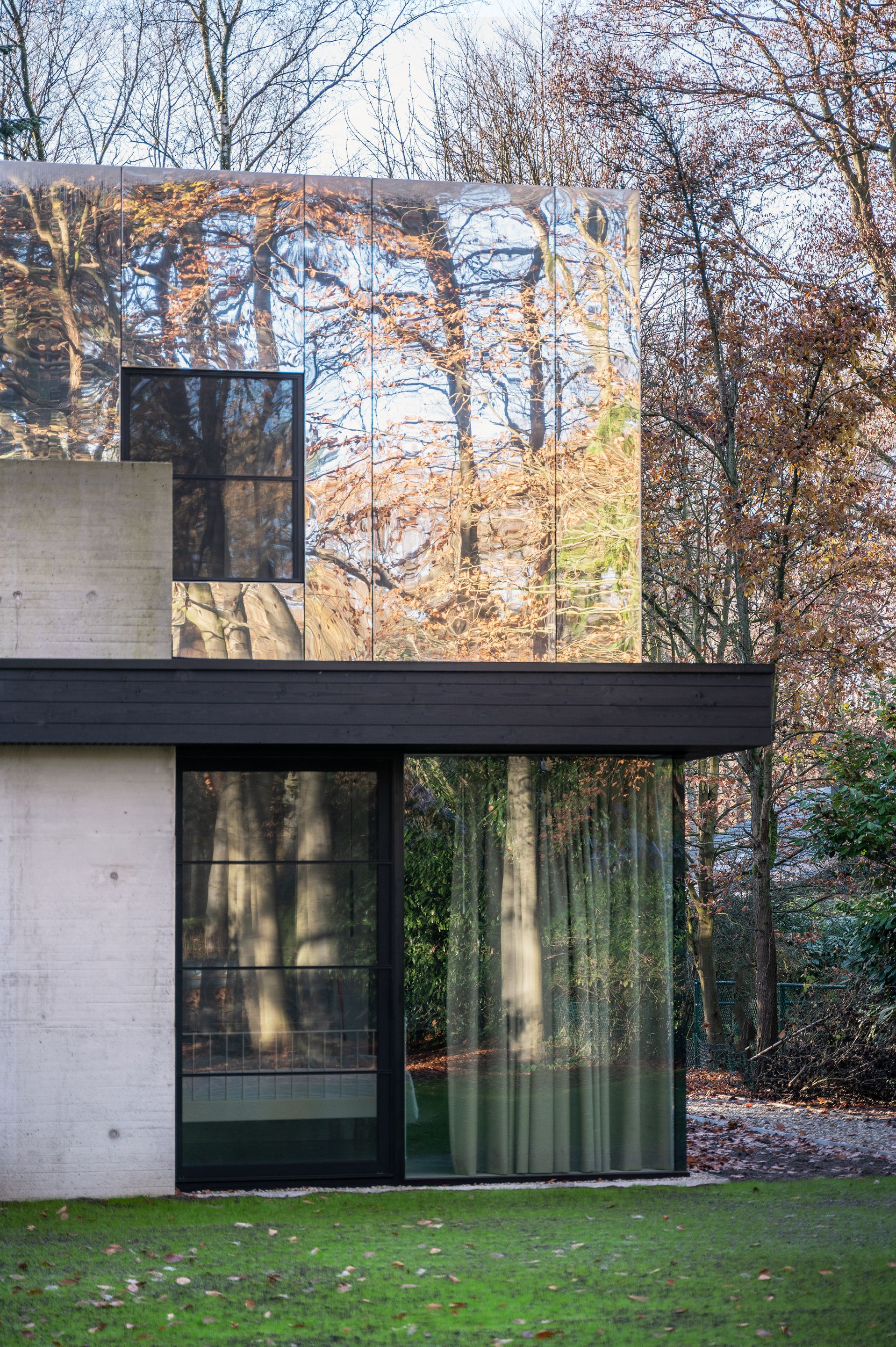Corner detail of mirrored upper volume reflecting bare branches above dark-framed sliding glass doors
