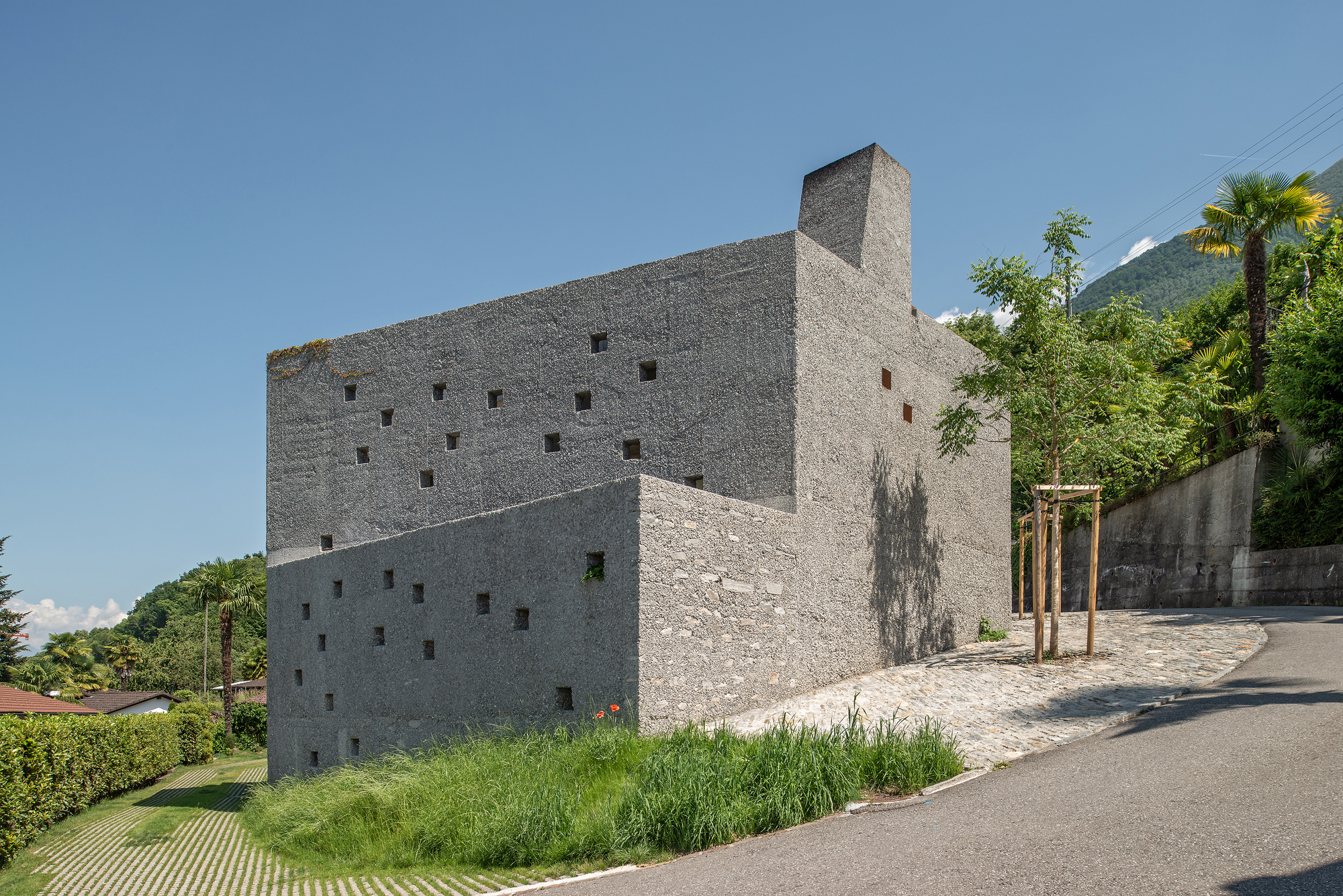Stacked concrete volumes with punched square windows on a sloping roadside with palm trees and mountains