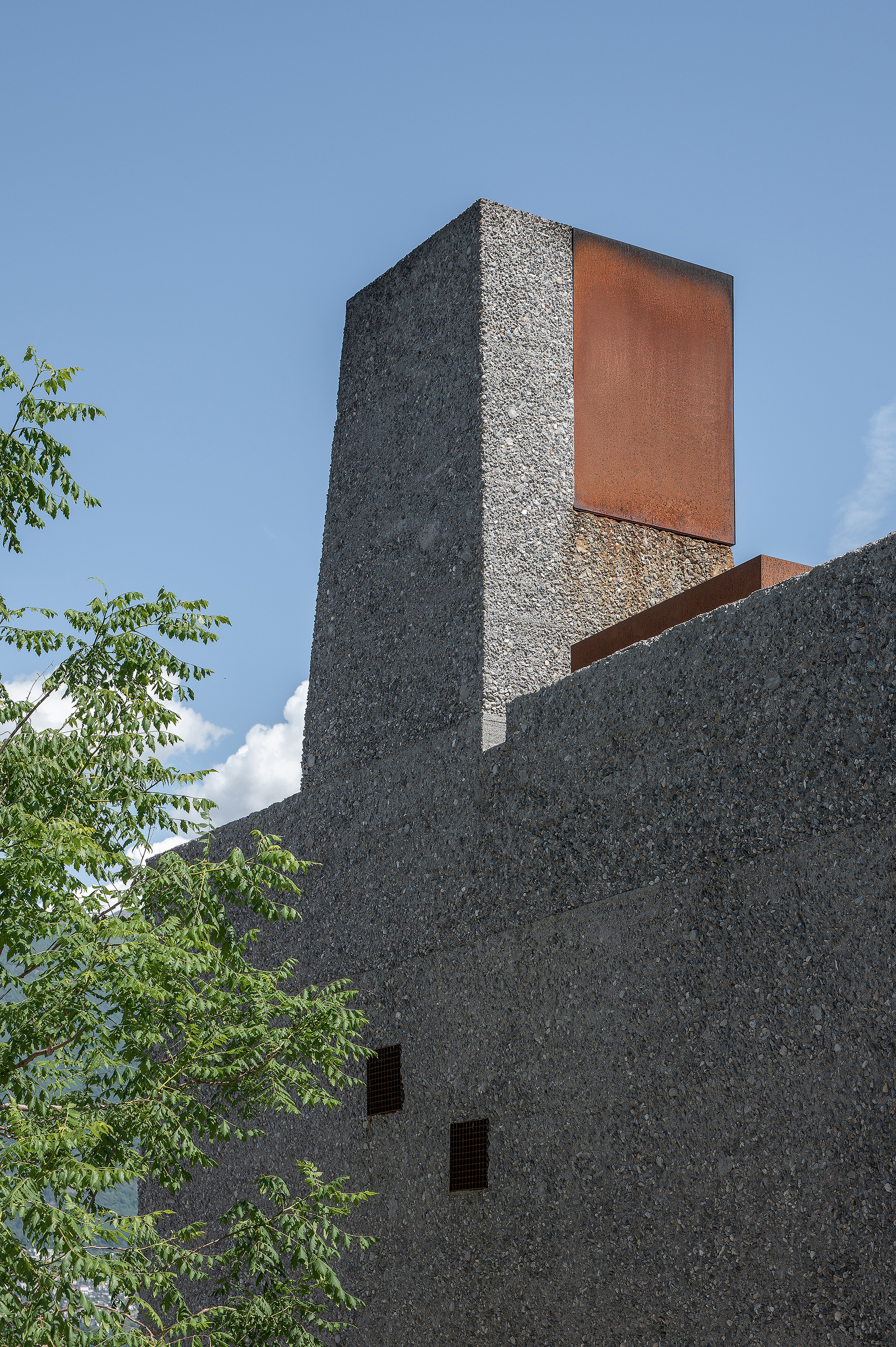 Corner chimney tower clad in weathered steel panels rising above textured aggregate concrete volumes