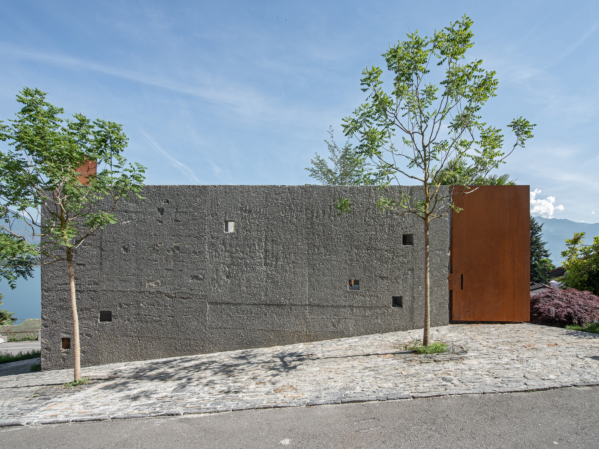 Street view of textured concrete facade with scattered windows and a weathered steel volume under a clear sky