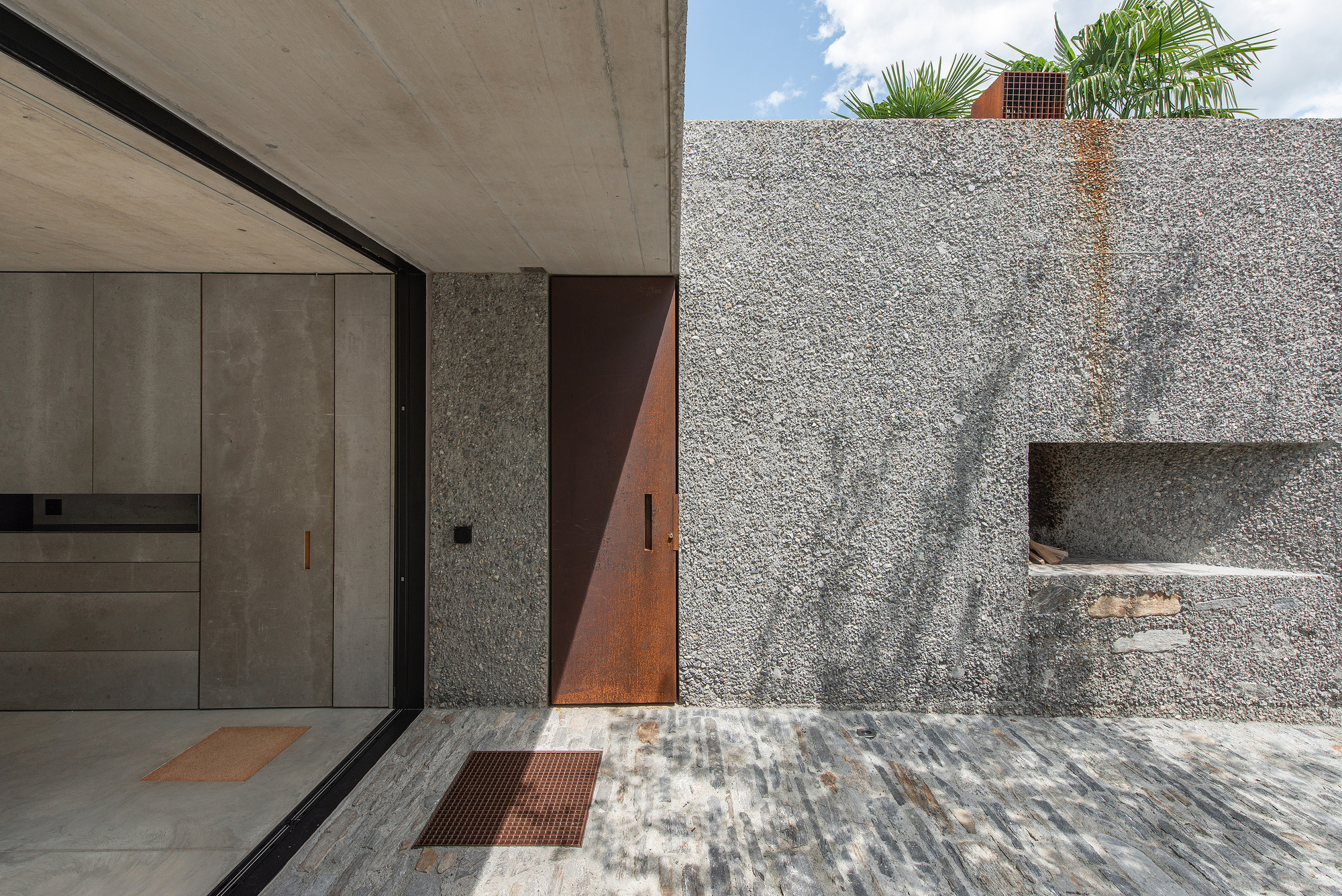 Covered entry passage with concrete soffit, large glazed opening, and weathered steel door in dappled sunlight