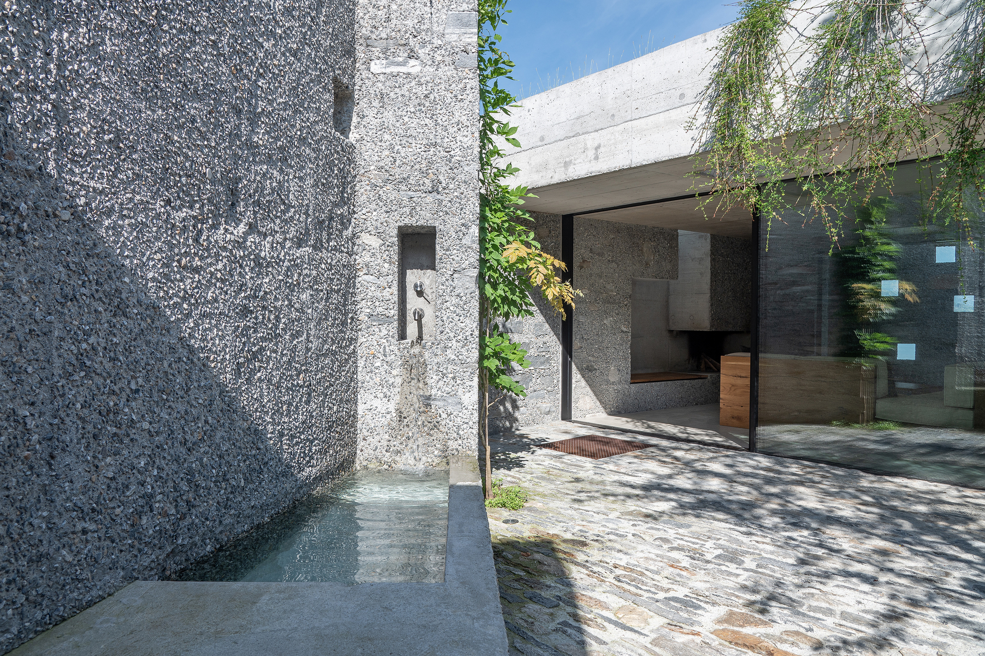Narrow entry corridor between textured concrete walls leading to glazed interior space with trailing vines overhead