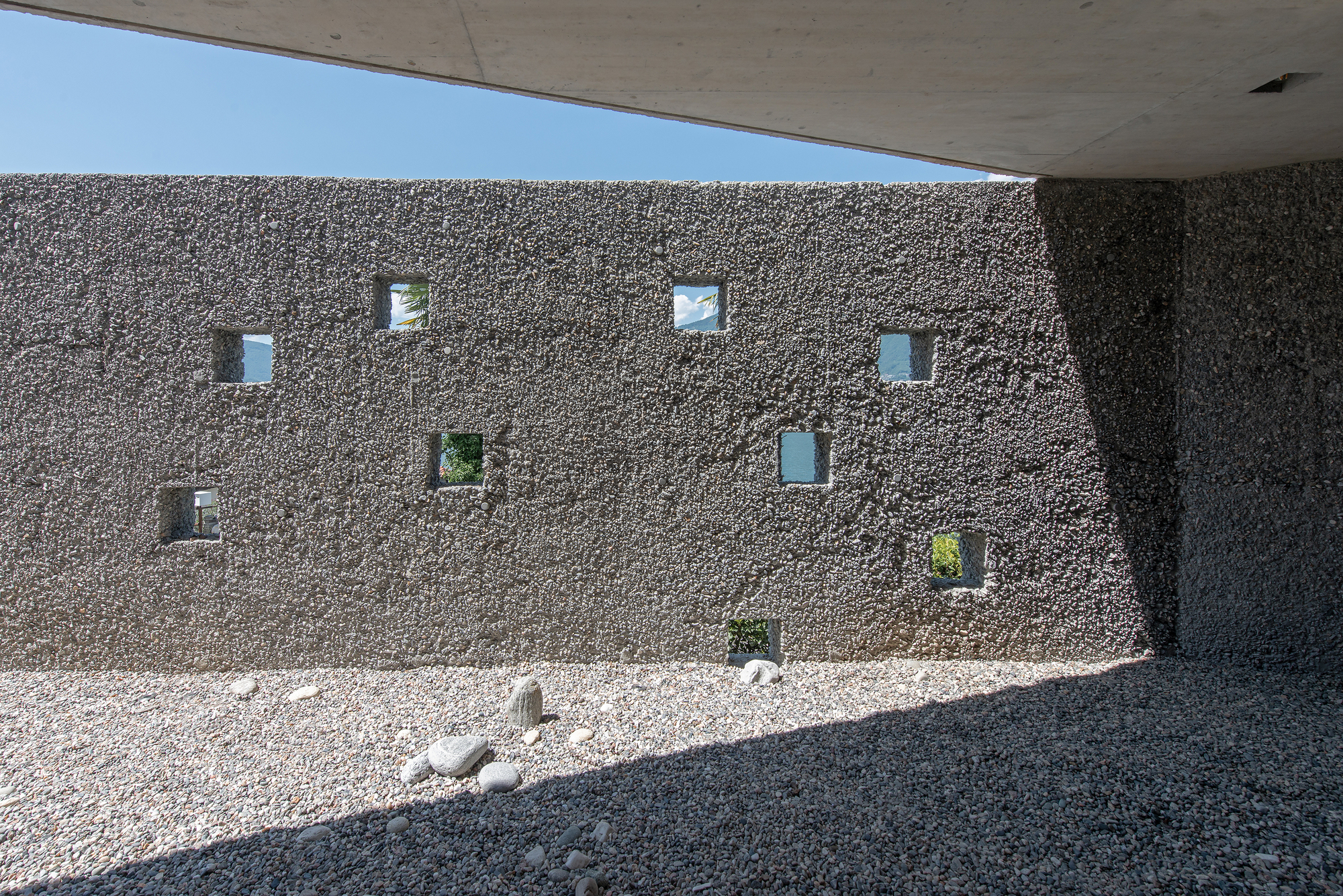 Exterior gravel courtyard wall with scattered square window openings beneath concrete overhang