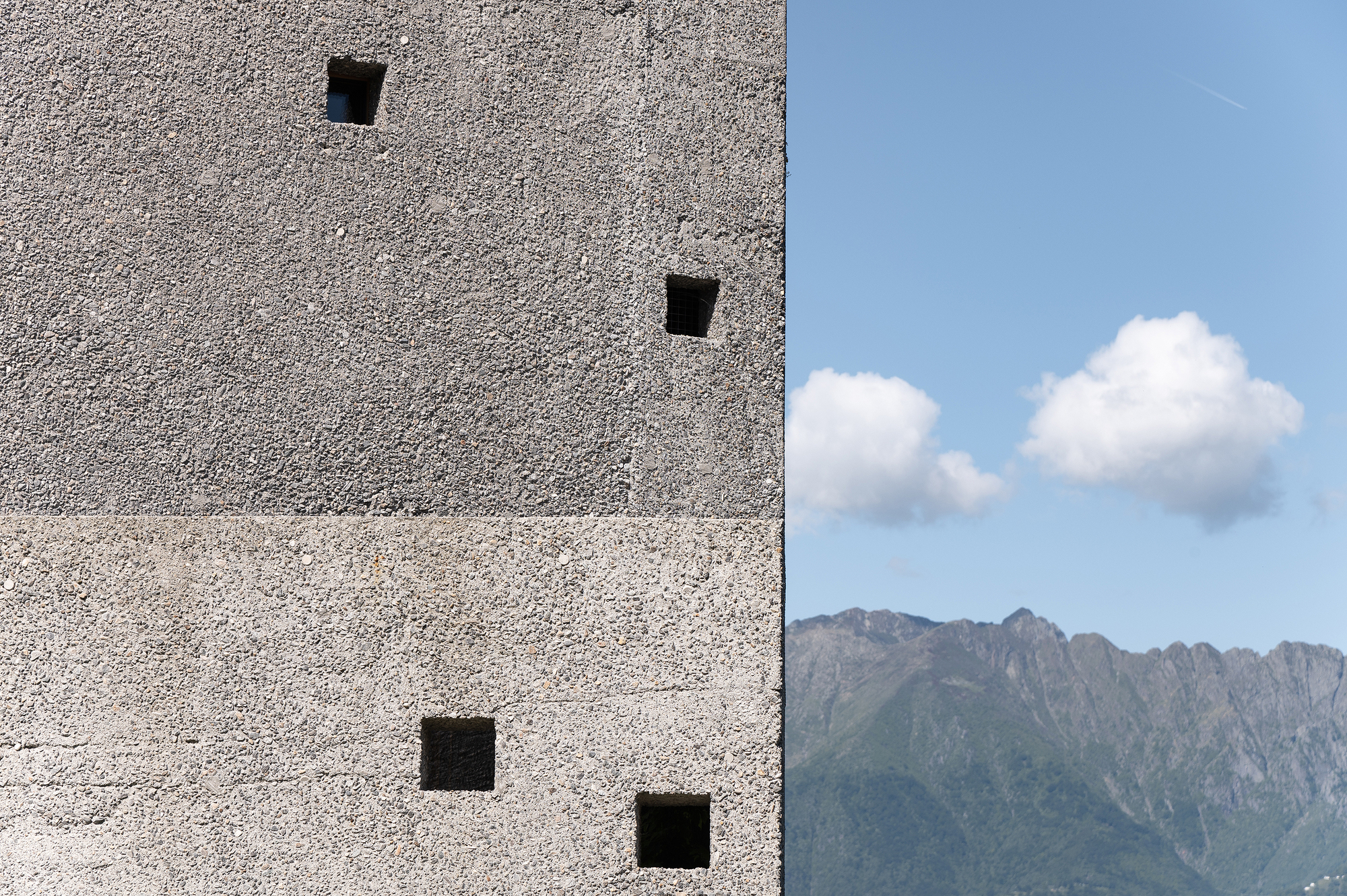 Textured aggregate concrete wall with punched square openings against distant mountain range and blue sky