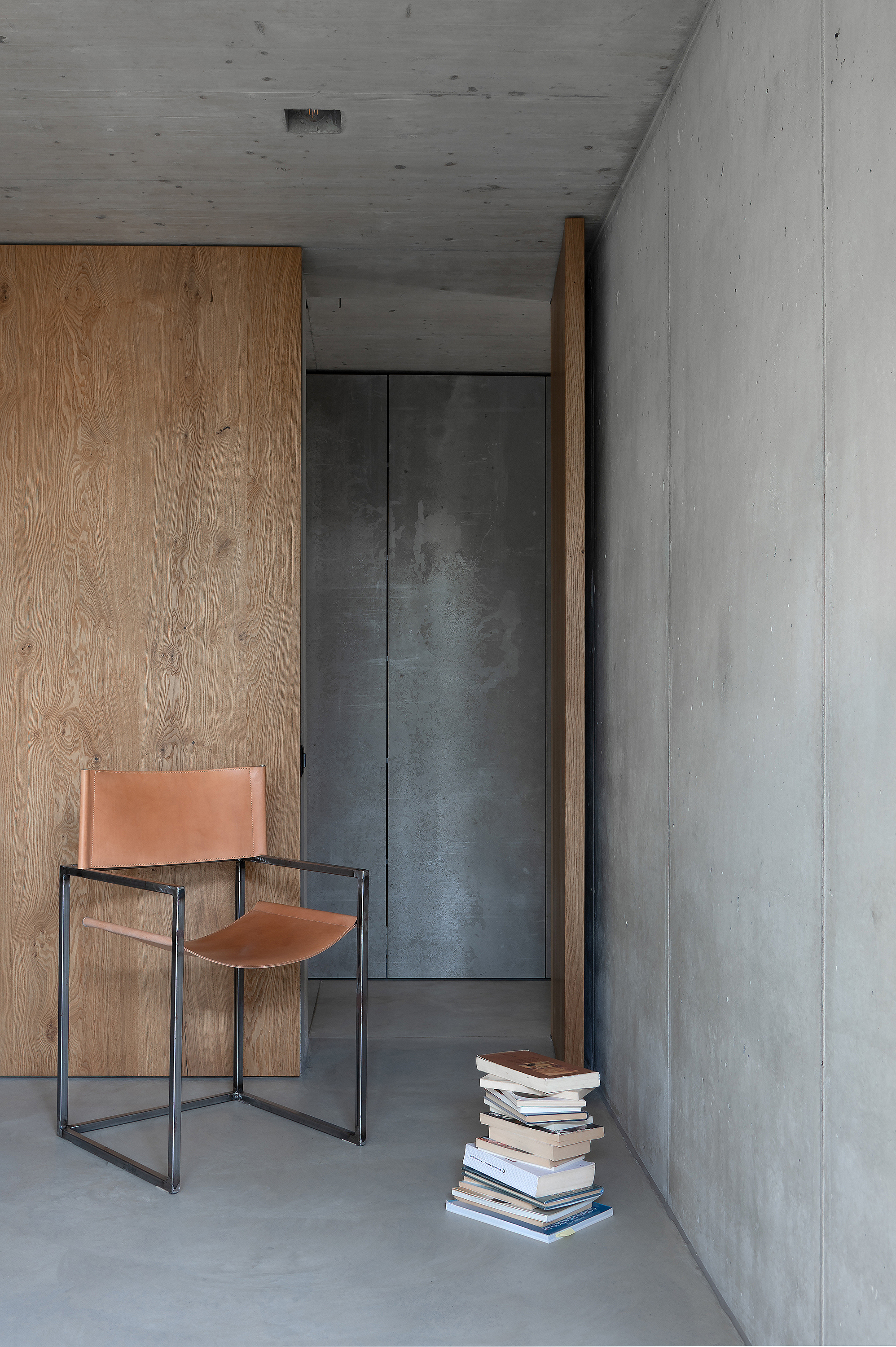 Interior corner with leather chair, stacked books on the floor, and timber cabinetry against concrete walls