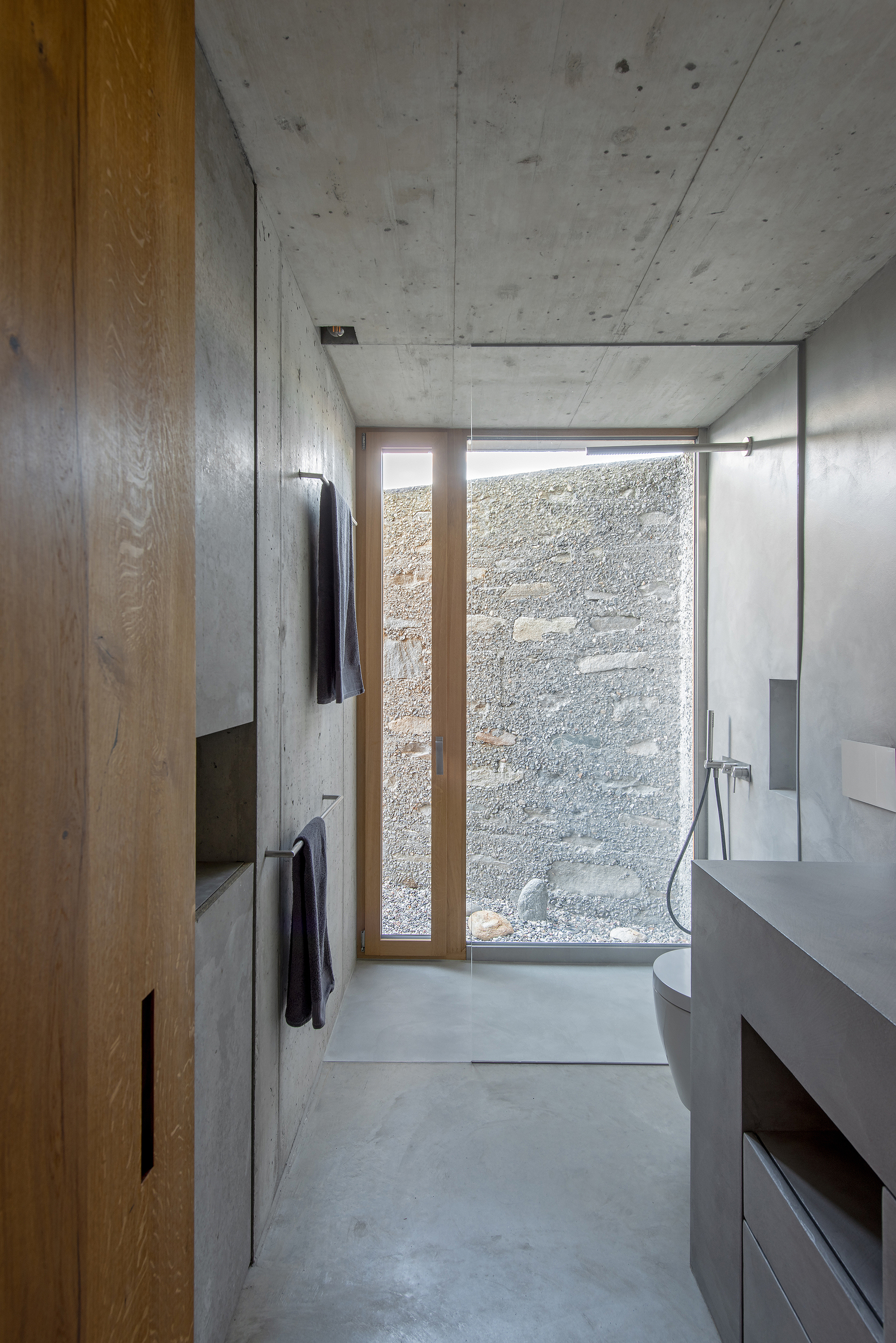 Bathroom with concrete walls, timber cabinetry, and a narrow window facing the textured courtyard wall