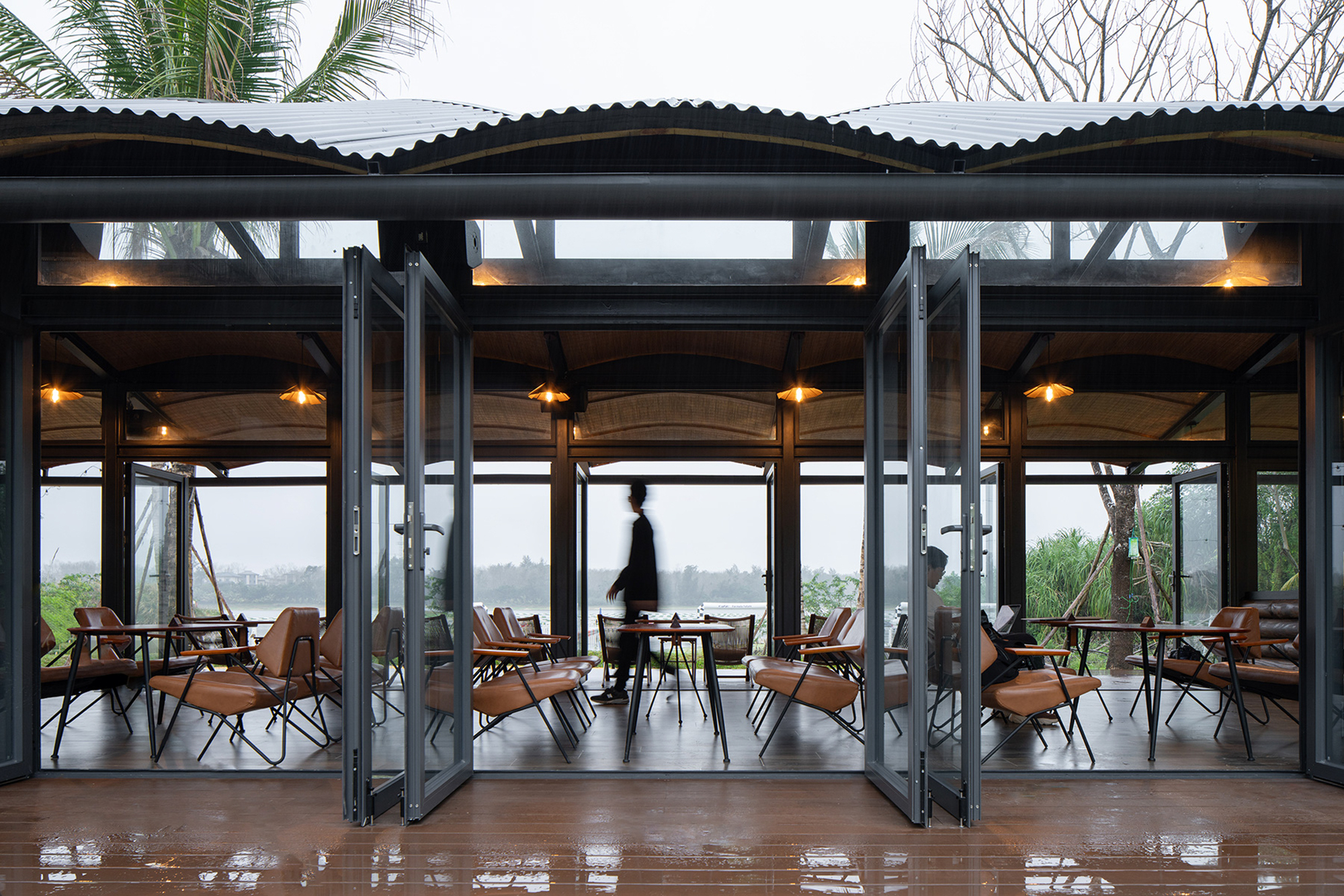 Open folding glass doors revealing the pavilion interior with woven rattan ceiling and a figure crossing the space