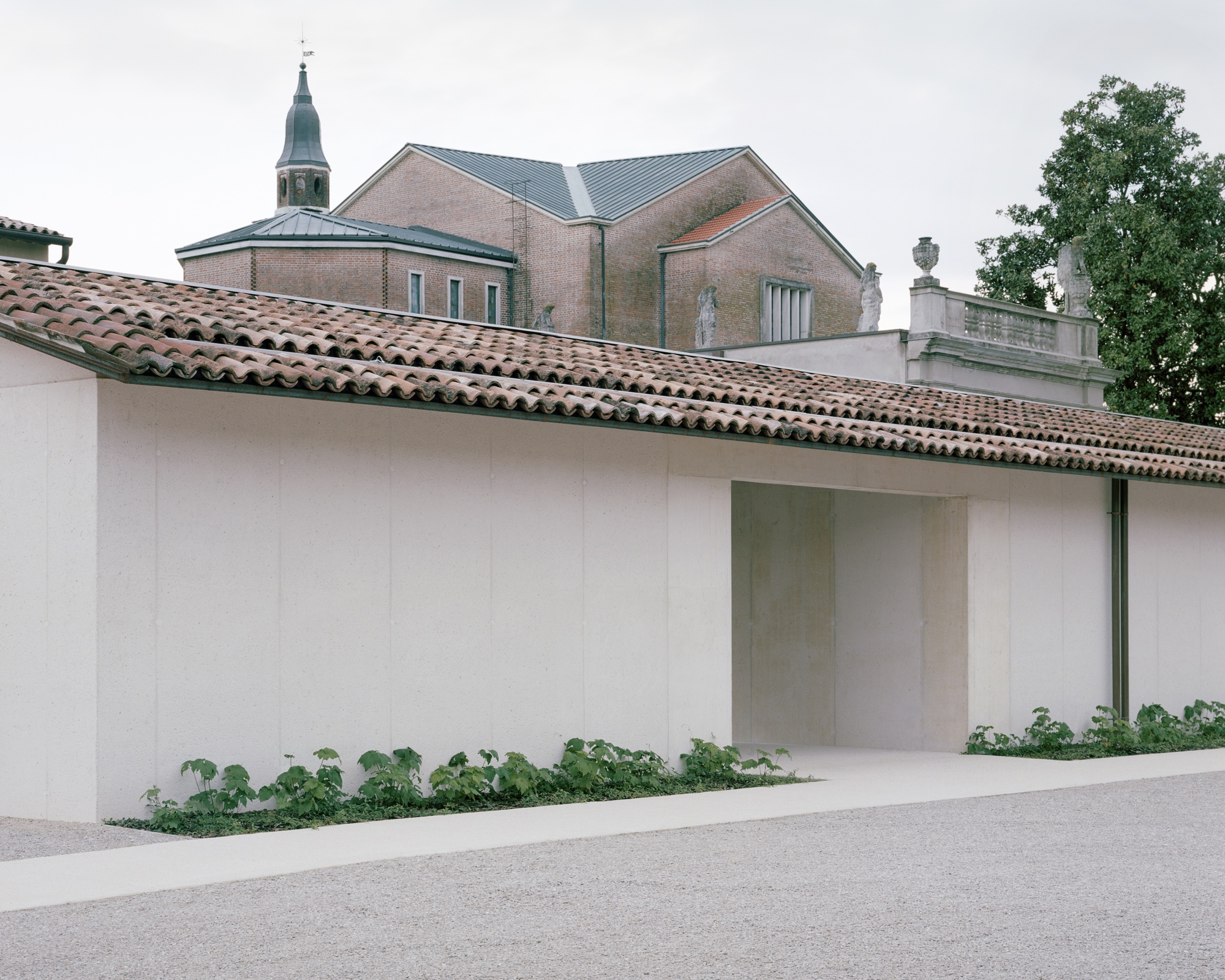 White plastered perimeter wall with tiled roof and recessed entry, with church campanile visible beyond