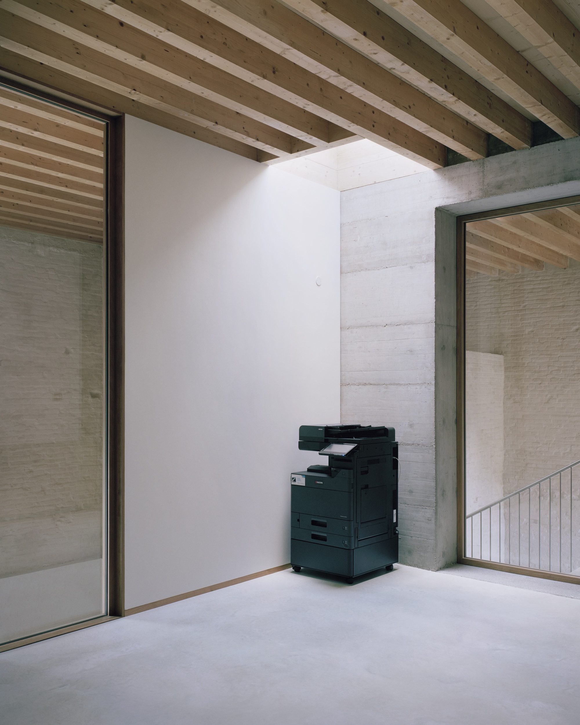 Corner nook with exposed timber joists overhead and printer stationed against white partition near glazed opening
