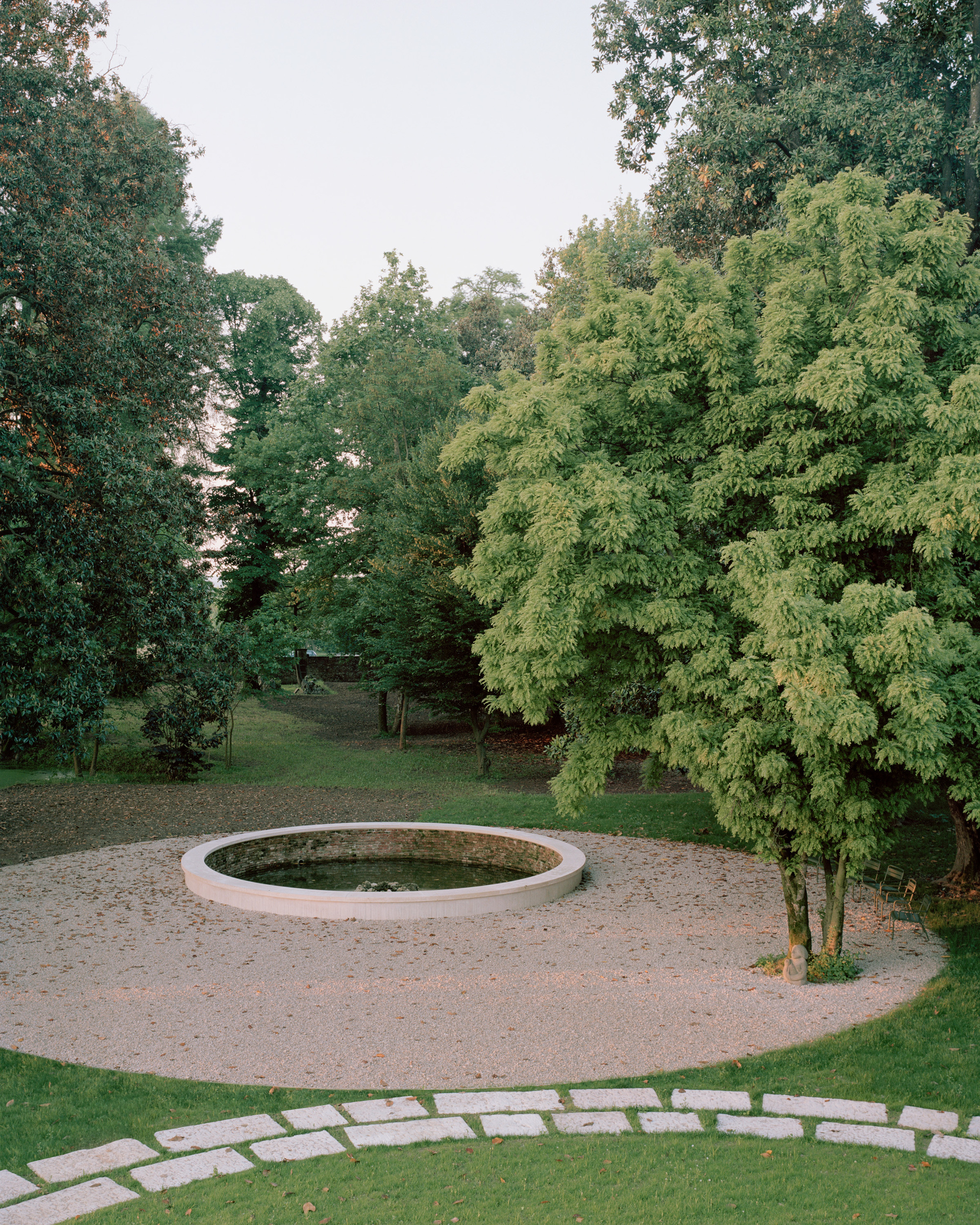 Circular stone fountain in a gravel courtyard surrounded by lawn and deciduous trees