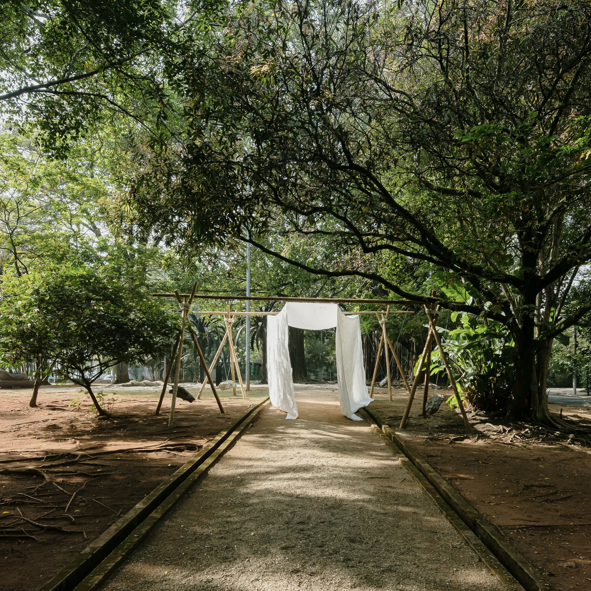 Timber frame pavilion with white fabric panels along a paved pathway beneath a canopy of trees