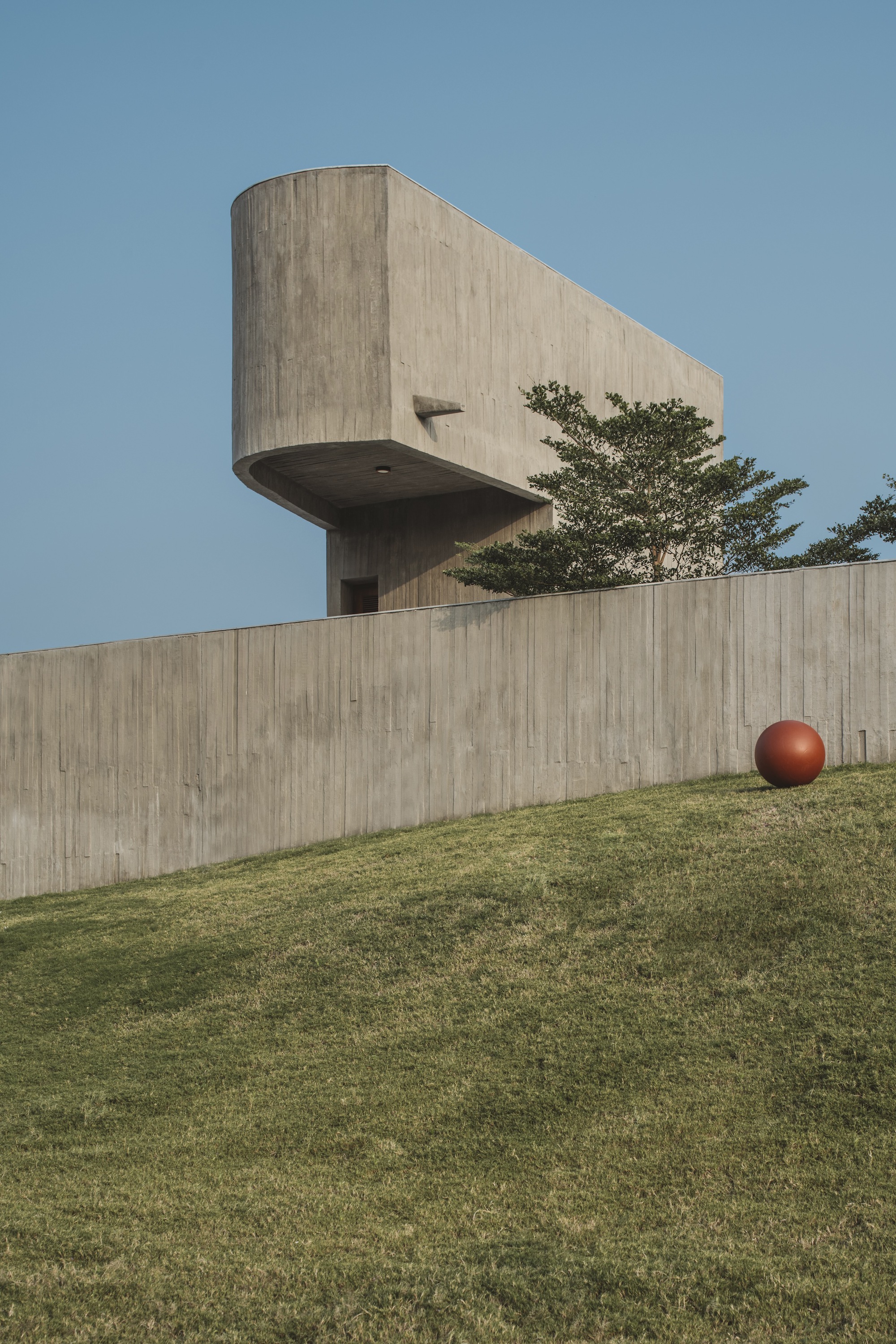 Board-formed concrete tower cantilevering over a curved retaining wall with a sphere on the grass