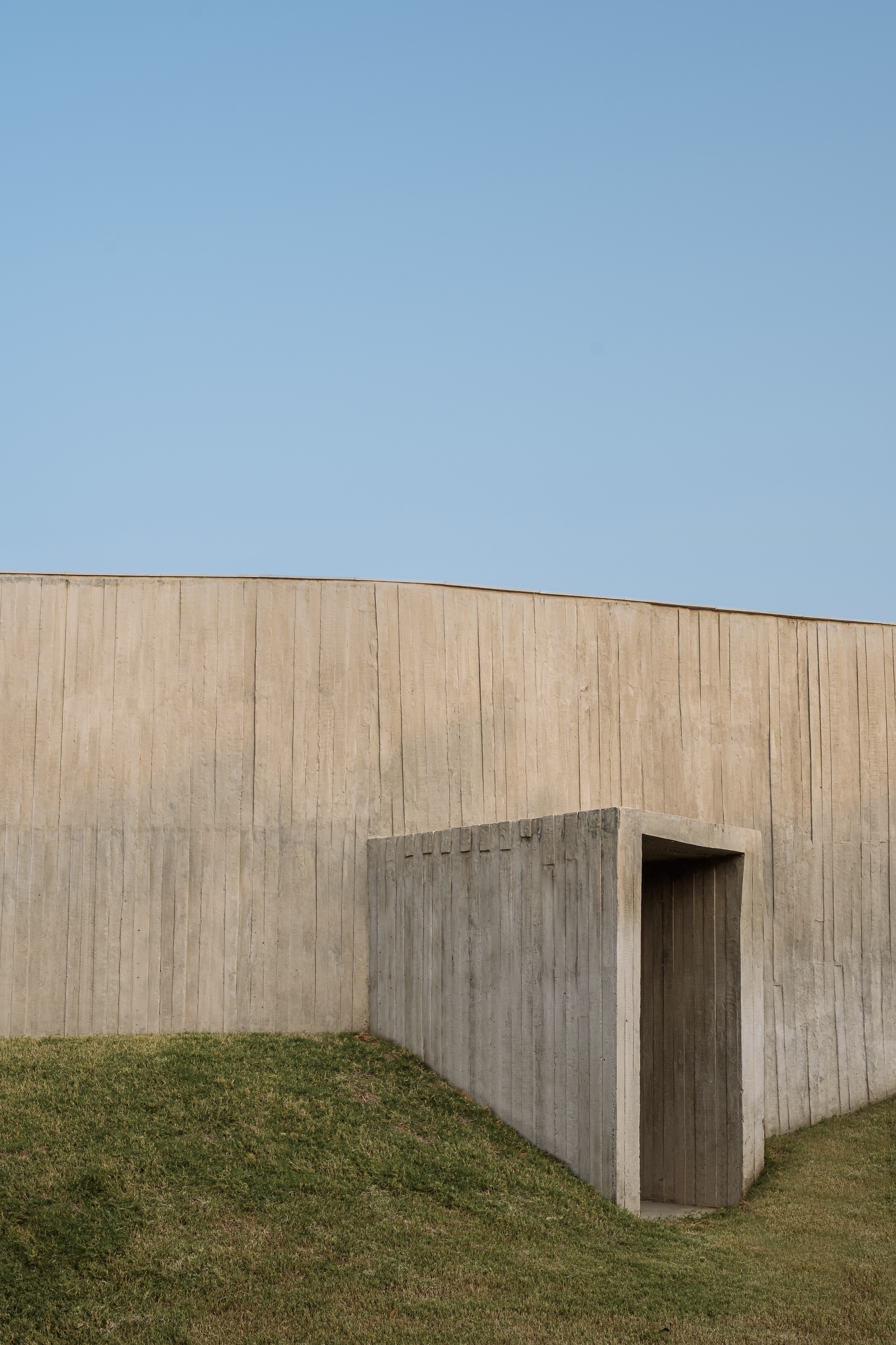 Board-formed concrete wall with recessed doorway threshold against a grass slope under clear sky