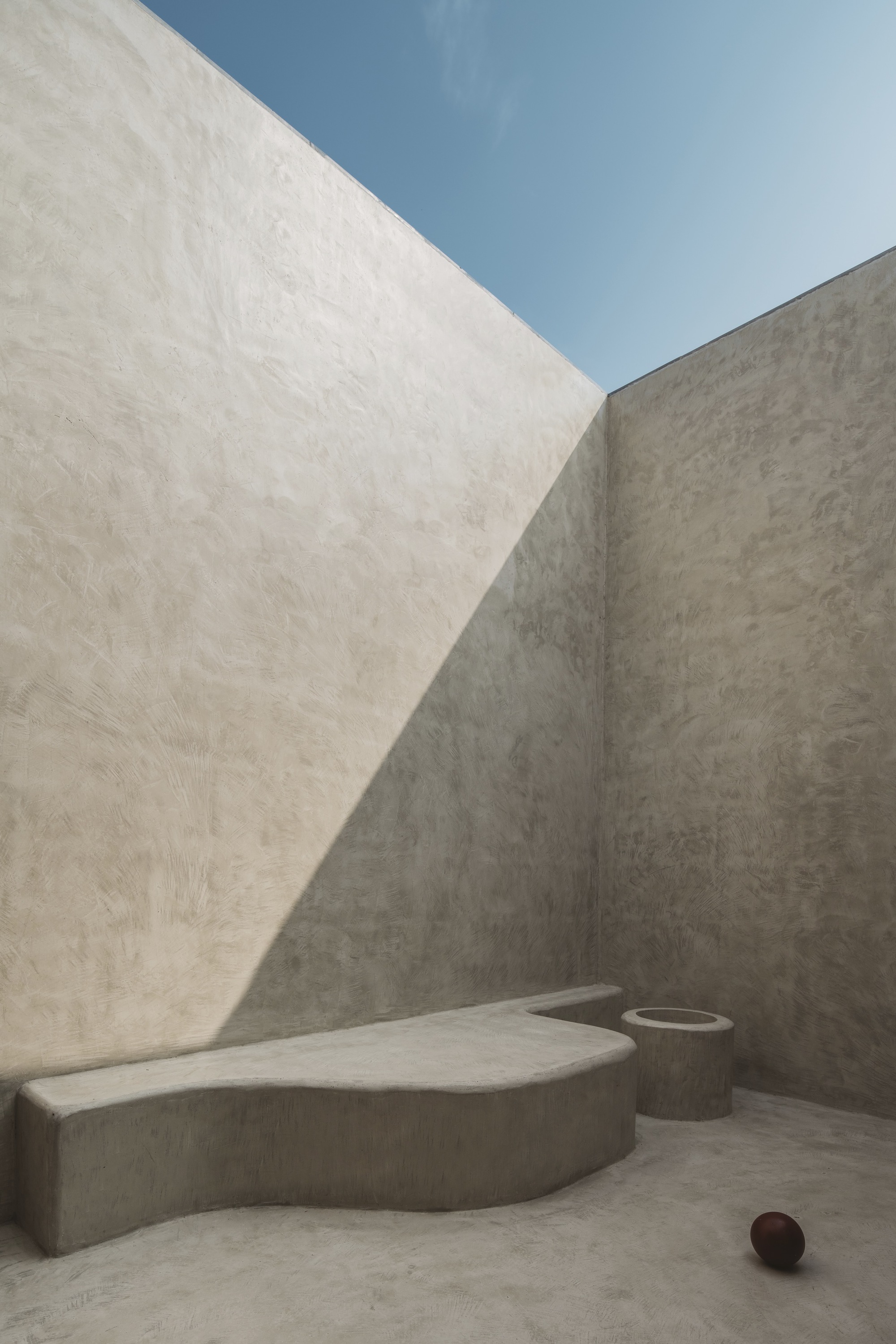 Concrete courtyard with angled walls, sculpted bench, cylindrical planter, and sharp diagonal shadows under open sky